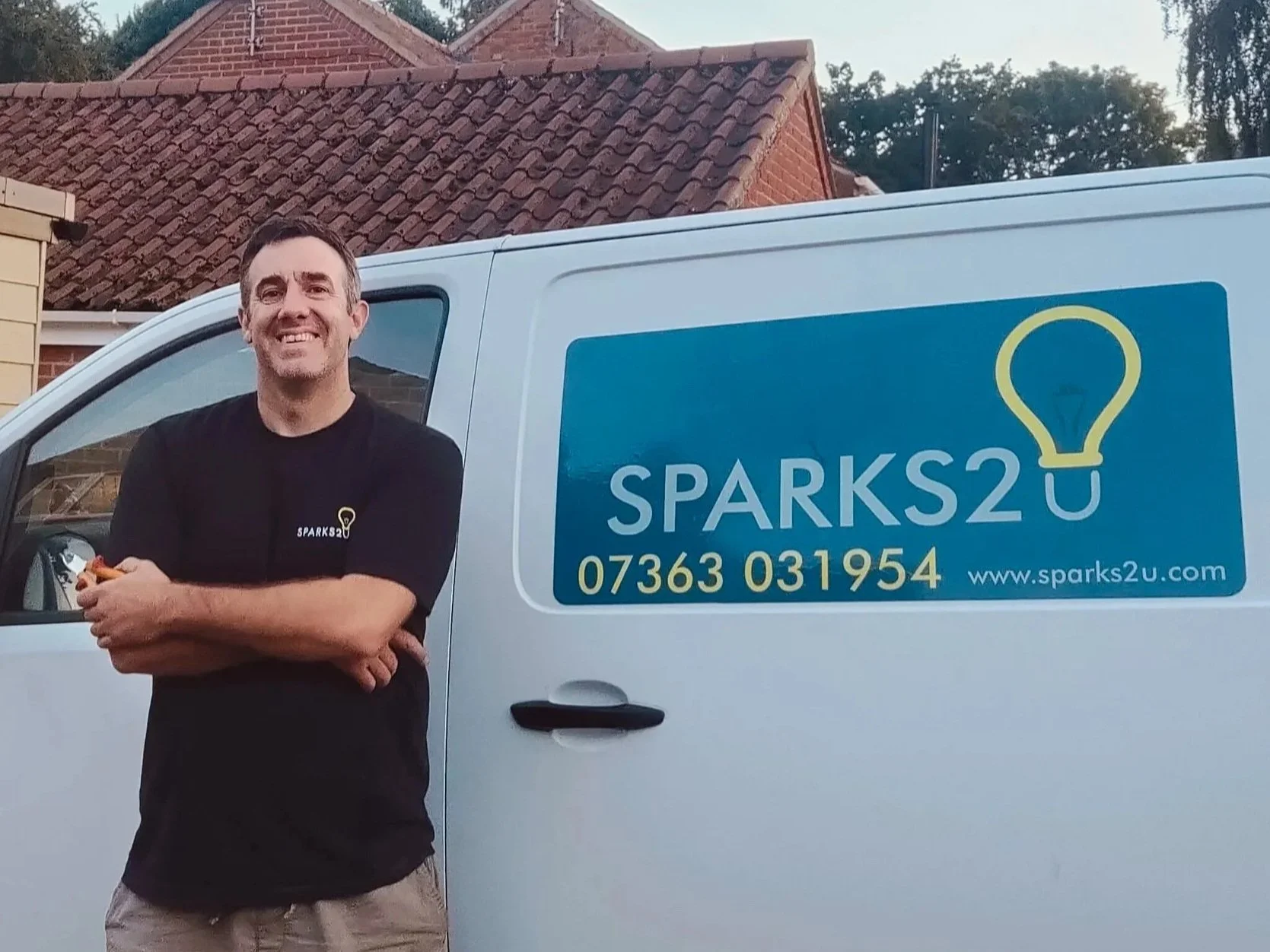 A man wearing a black t-shirt with a yellow lightbulb and 'SPARKS2U' logo, smiling and holding a small orange object, standing next to a white work van with the same logo, phone number, and website displayed on the side.