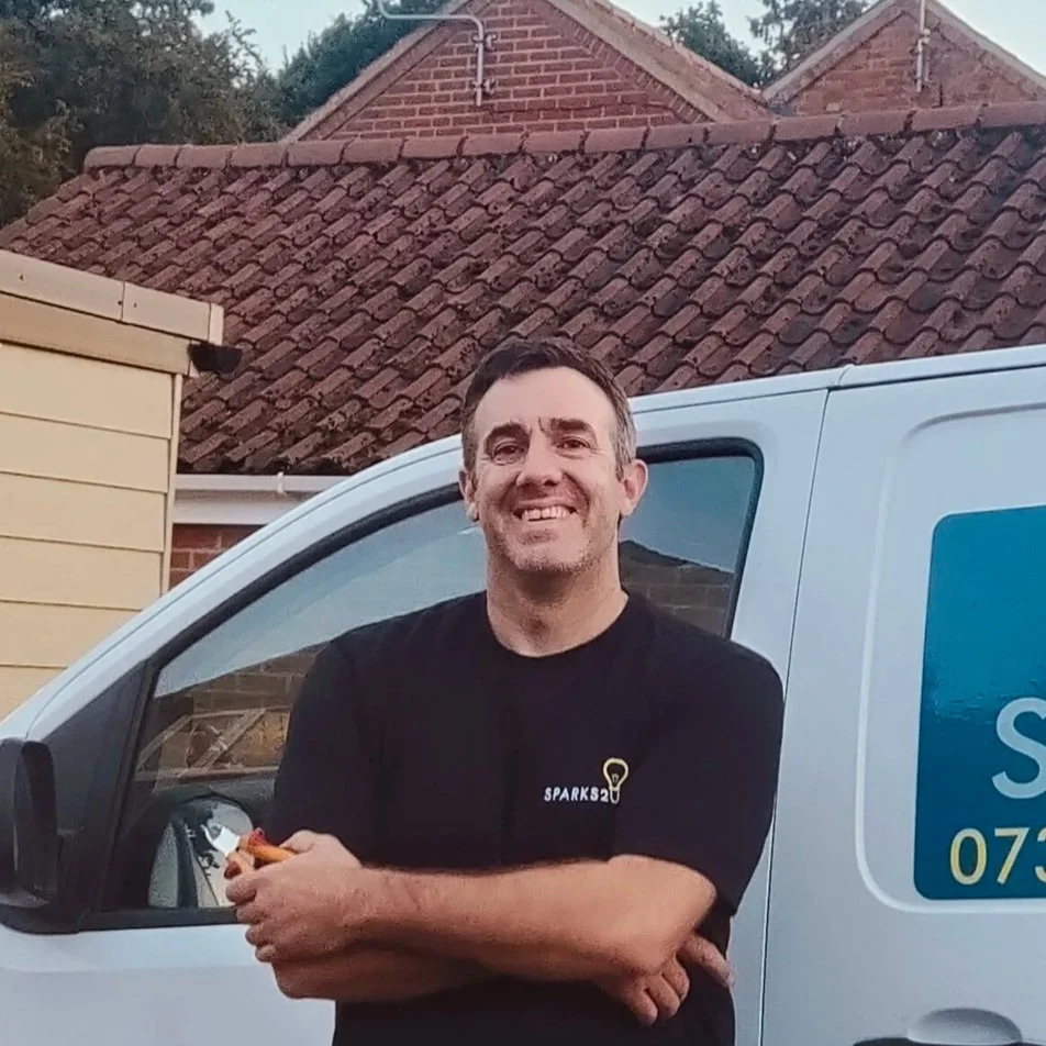 A man smiling and standing outdoors in front of a white service van with partially visible blue and yellow logo and service contact, with houses and a tiled roof in the background.