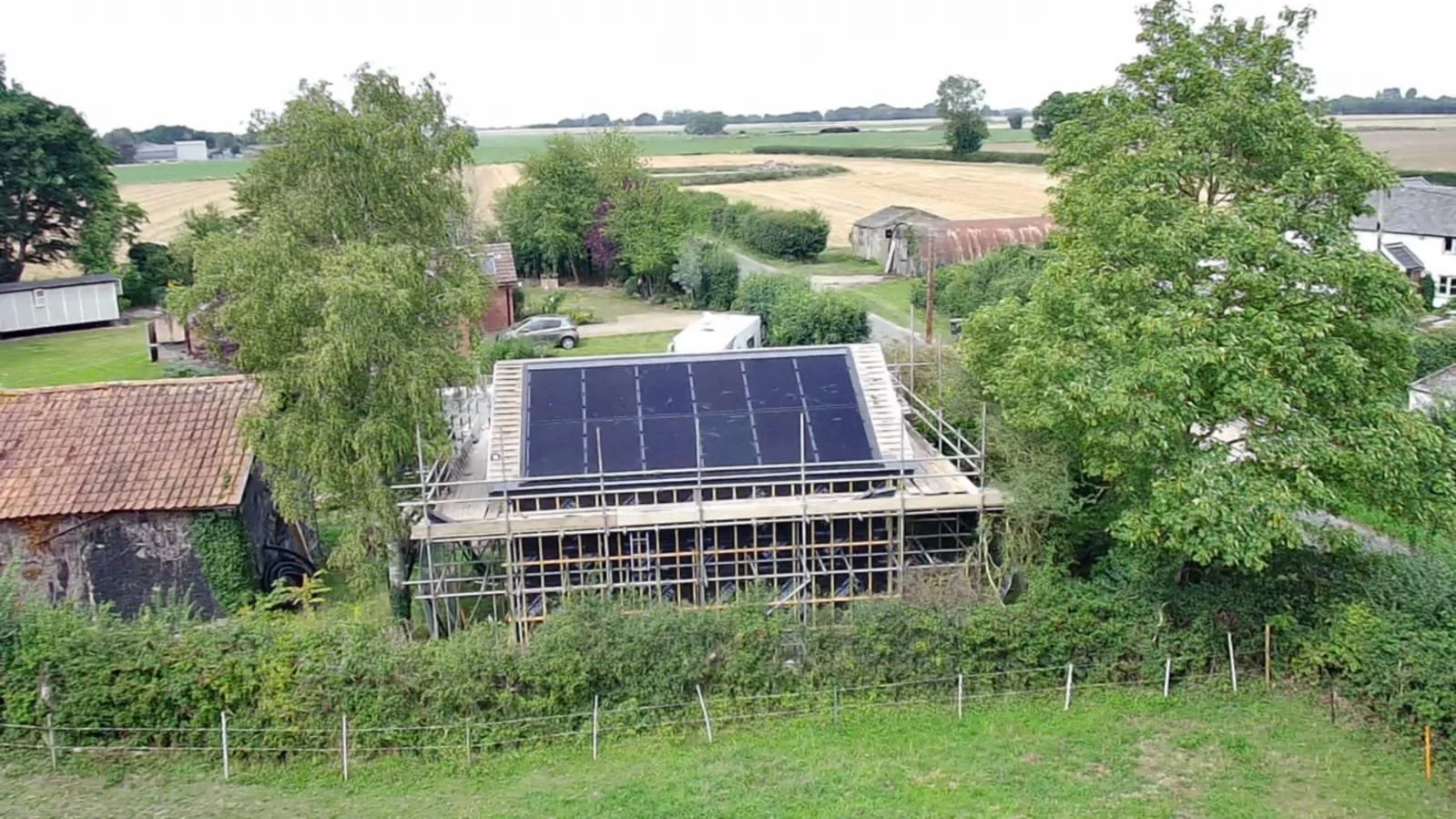 A house under construction with solar panels on the roof, surrounded by trees, in a rural area with fields and farmland in the background.