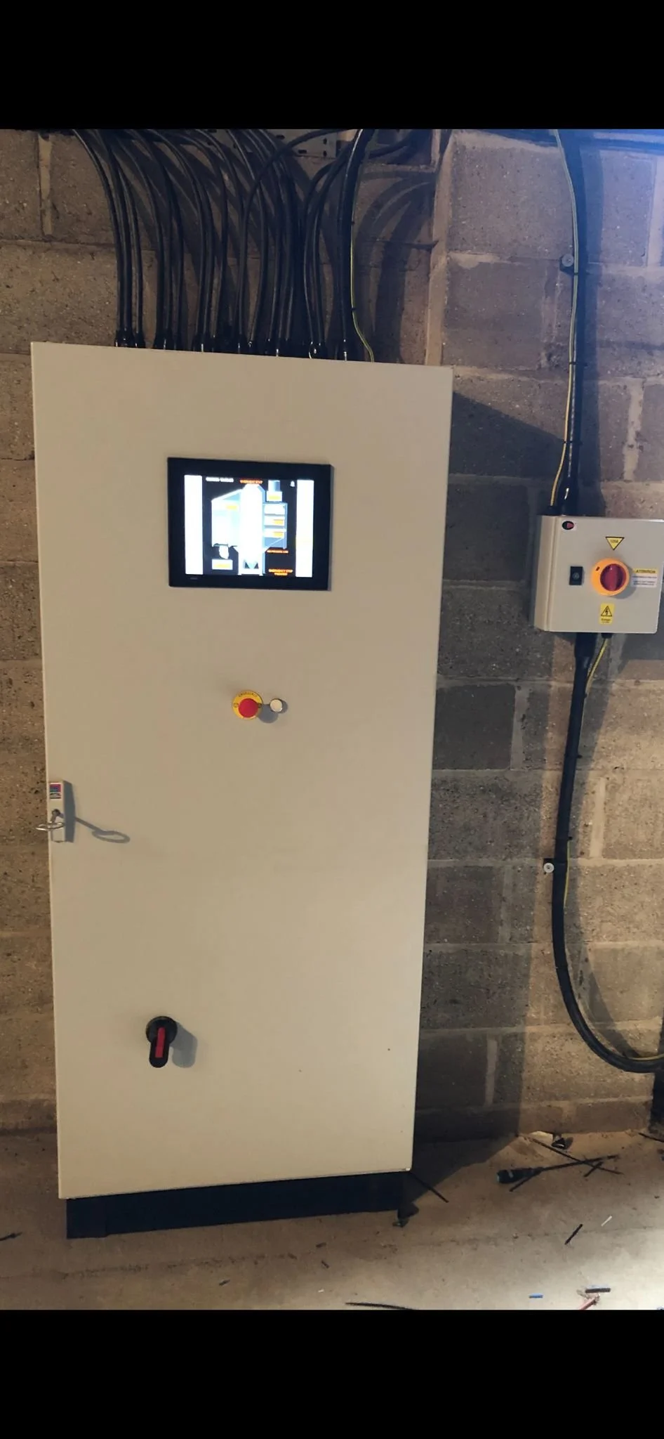 Industrial electrical control panel mounted on a wall with wires connected at the top, a small screen displaying system information, and emergency stop buttons.