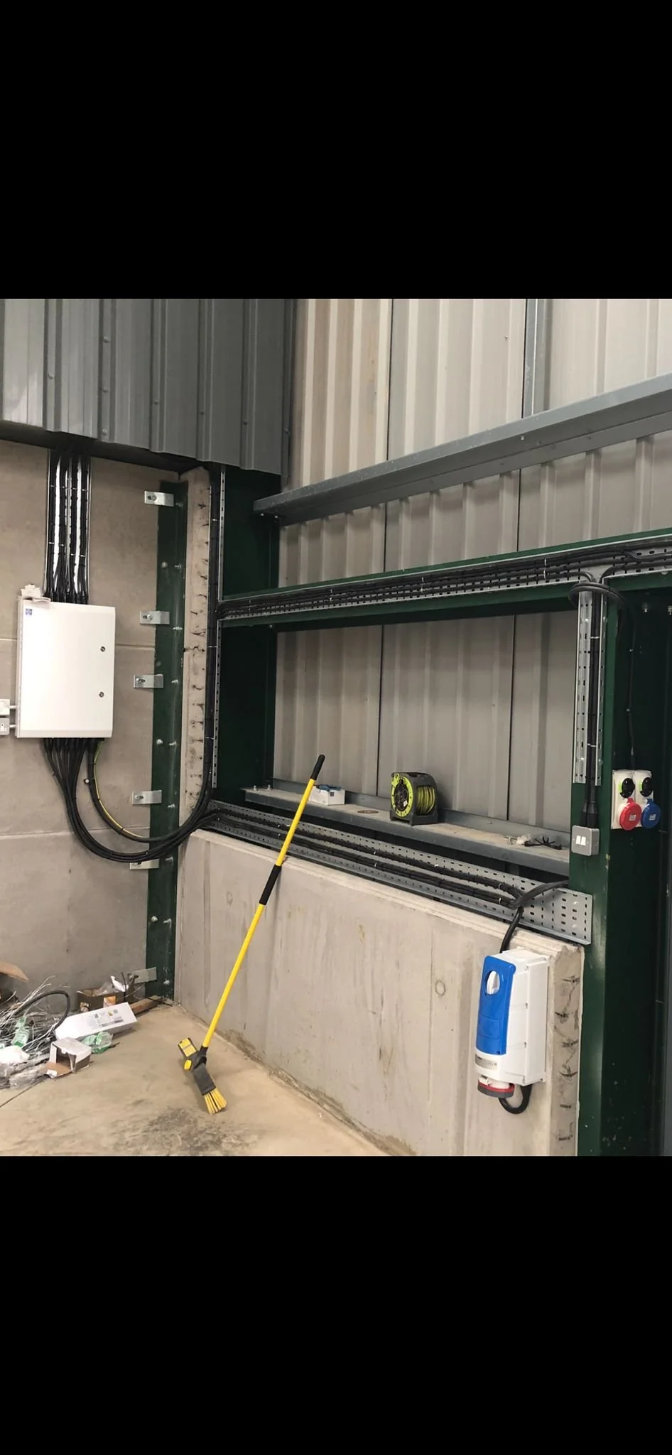 An industrial workspace shows a shelving unit with electrical wiring and components, a yellow broom leaning against the concrete wall, and tools and equipment on the floor and shelves.
