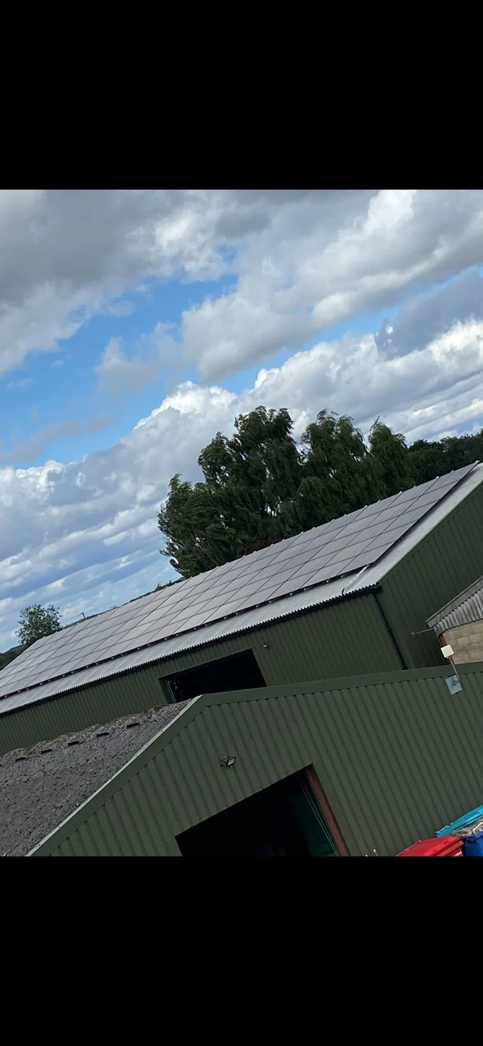 A large building with green siding and a metal roof equipped with solar panels, situated outdoors under a partly cloudy sky with trees in the background.