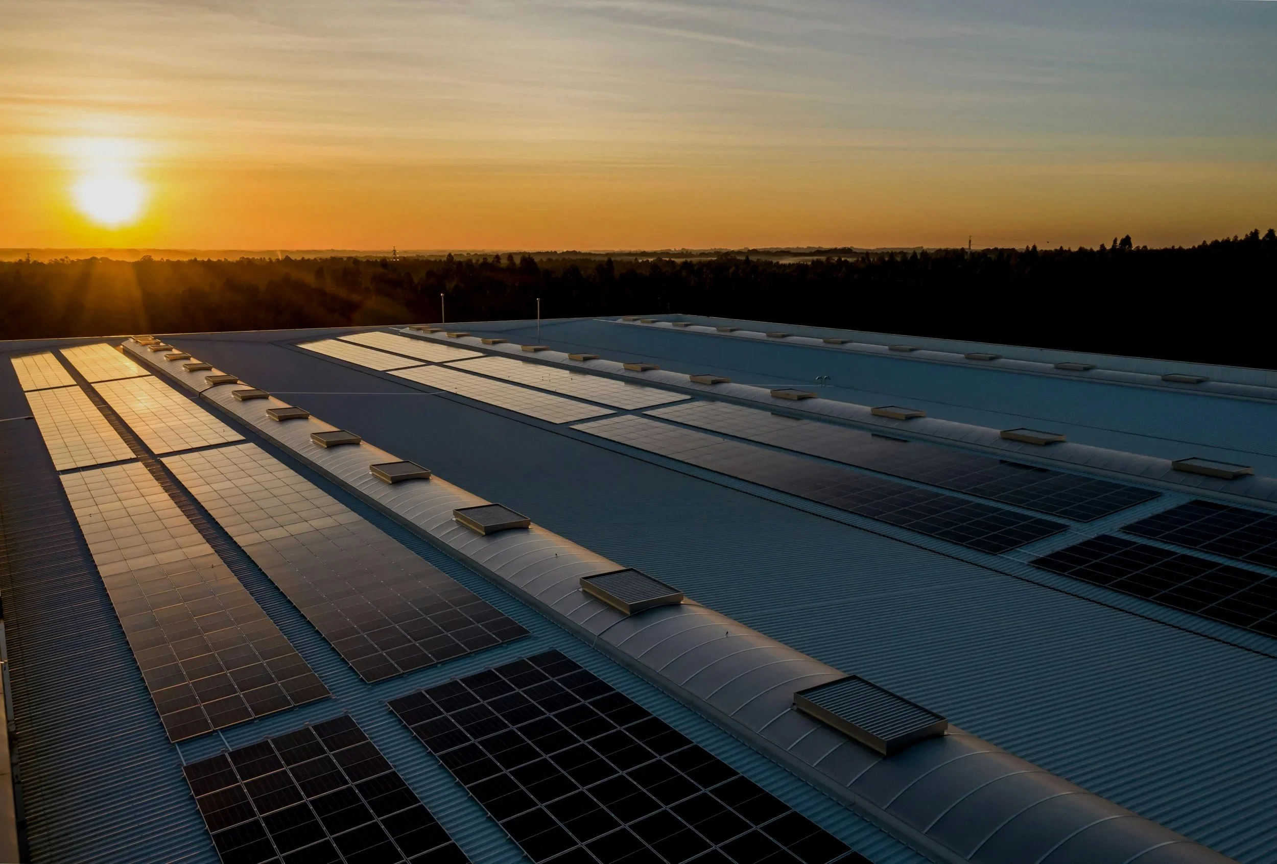 Sunset over a large metal roof with solar panels installed on it, with a distant tree line on the horizon.