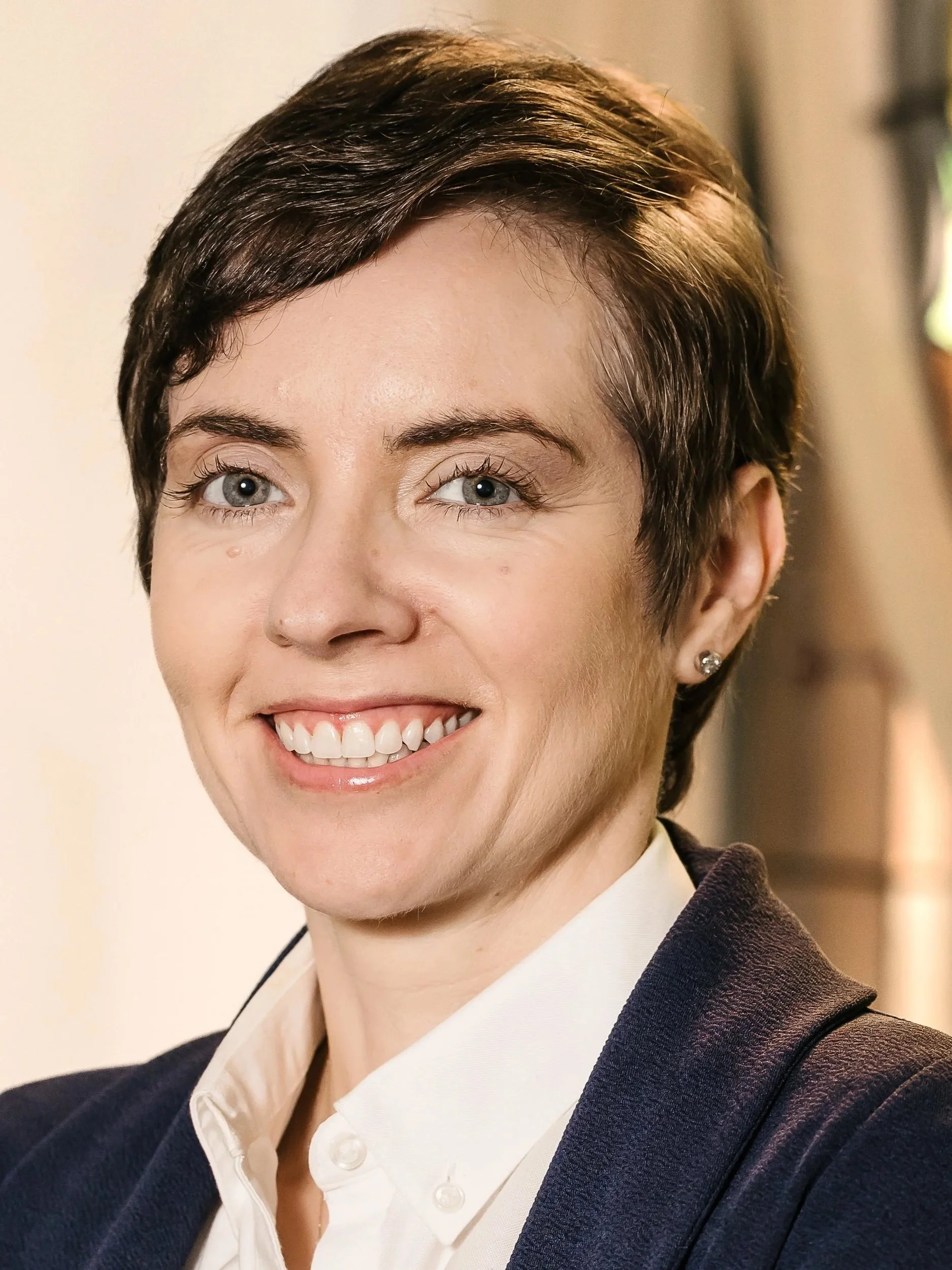 Smiling person with short hair wearing earrings and a dark top, with a blurred background.