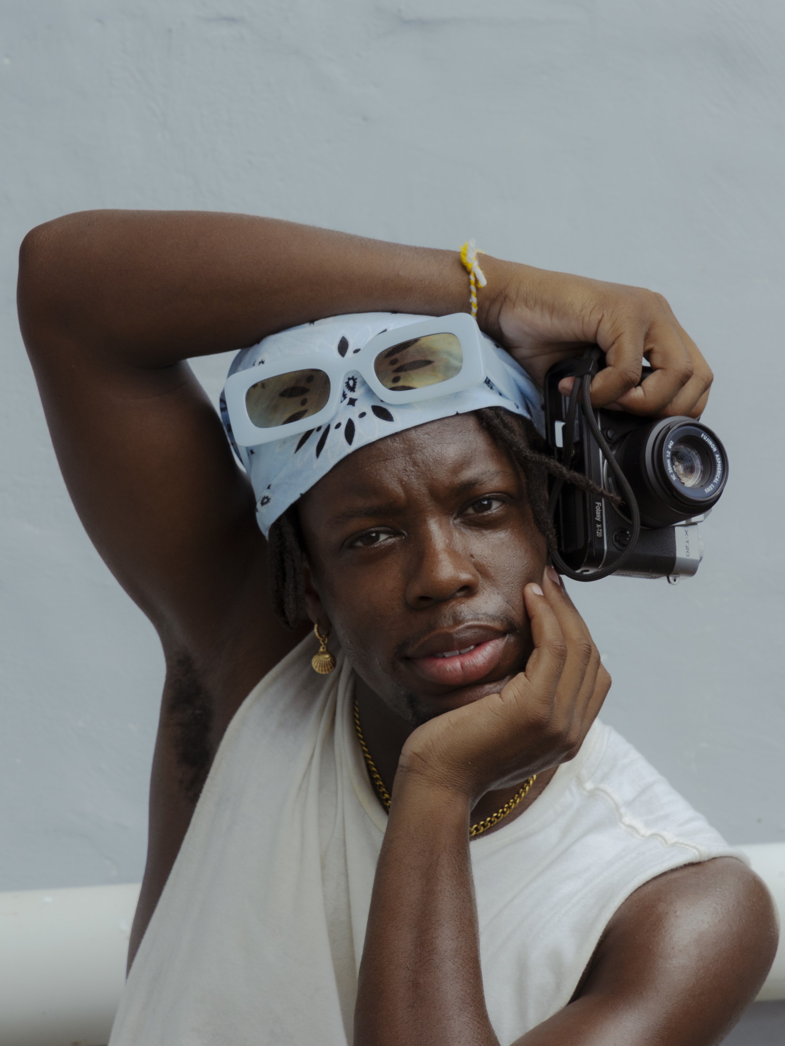 A person with medium brown skin and wearing a white sleeveless shirt, a white bandana with black patterns, yellow earrings, and a gold necklace, is holding a camera to their right ear and resting their chin on their left hand. The person's face is partially visible, and their eyes are looking towards the camera. The background is plain and light-colored.