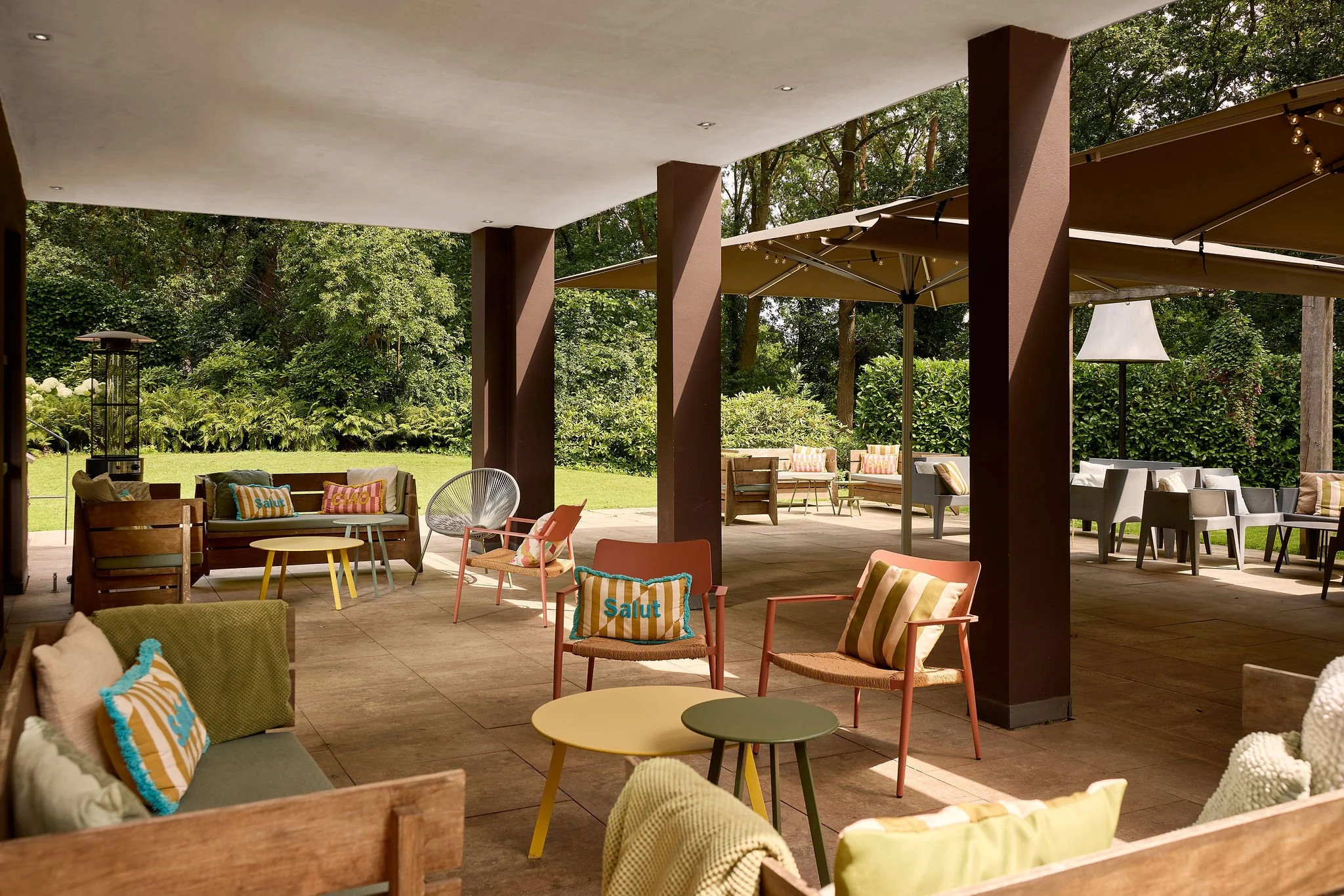 Je ziet kussens, tafels en stoelen. Leuke lounge sets. Veel groen en een overkapping