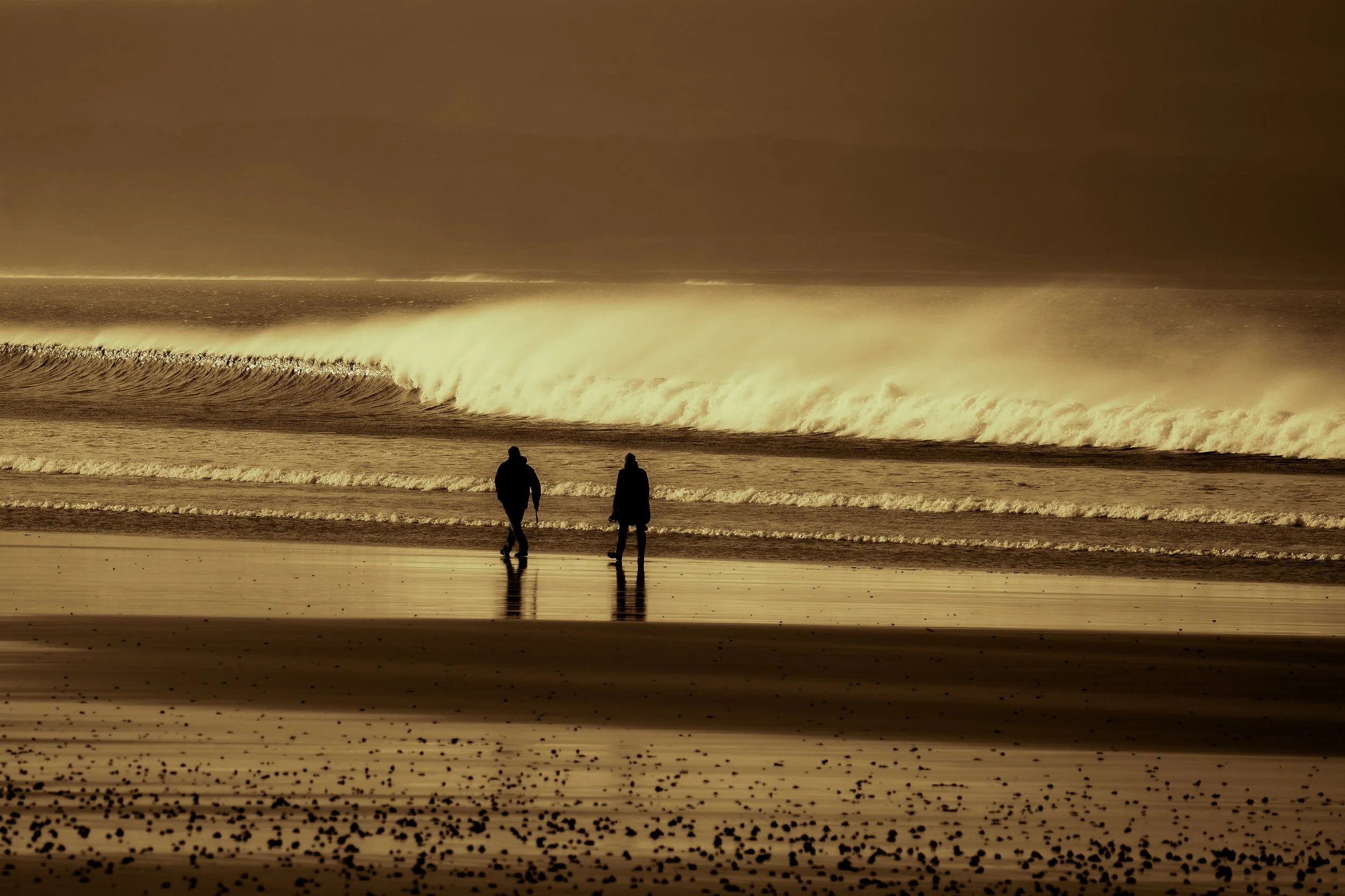 Two silhouetted people walking along a beach with large ocean waves crashing in the background under a cloudy sky with a sepia tone