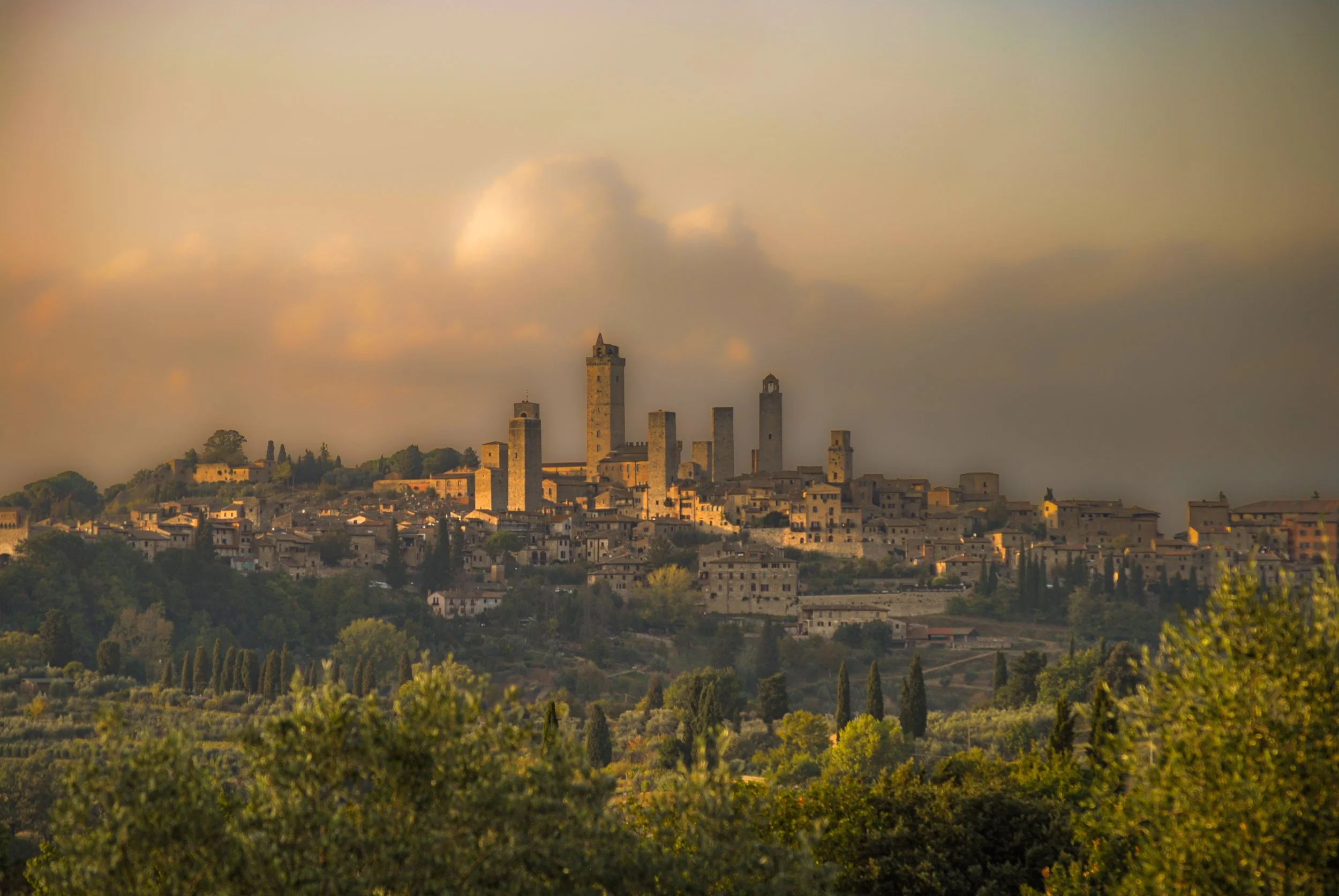 San Gimignano in Tuscany, Italy, photographed at golden hour with medieval stone towers, olive trees, cypress trees, and the warm Tuscan countryside surrounding the hill town.