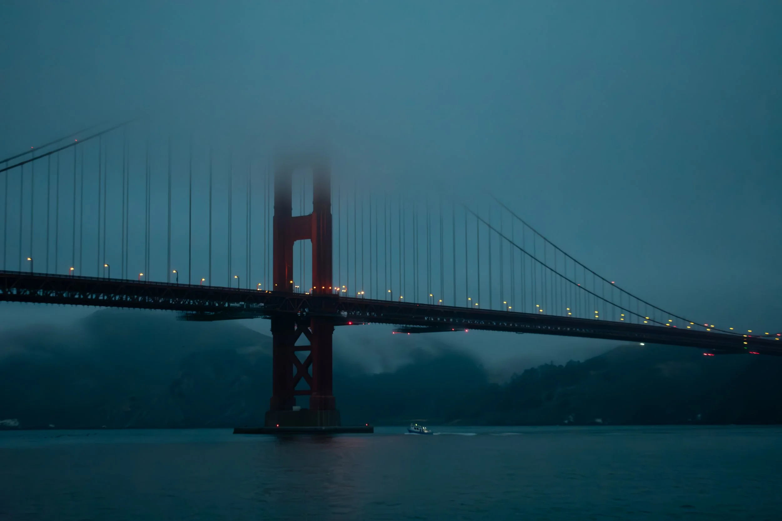 Golden Gate Bridge shrouded in fog at dusk, with a boat on the water below.