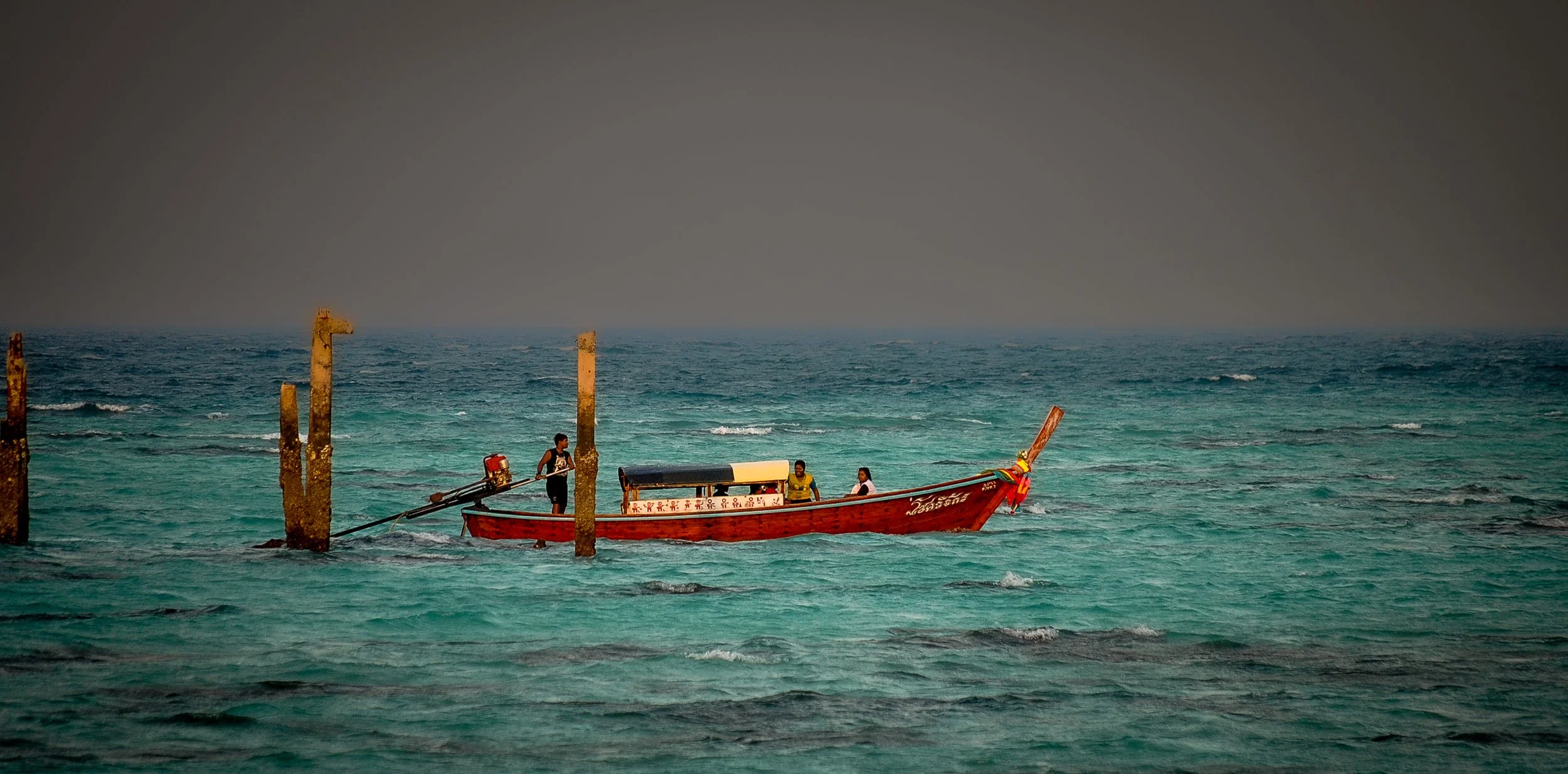 Small wooden boat with four people, navigating near rusted wooden posts in turquoise ocean water