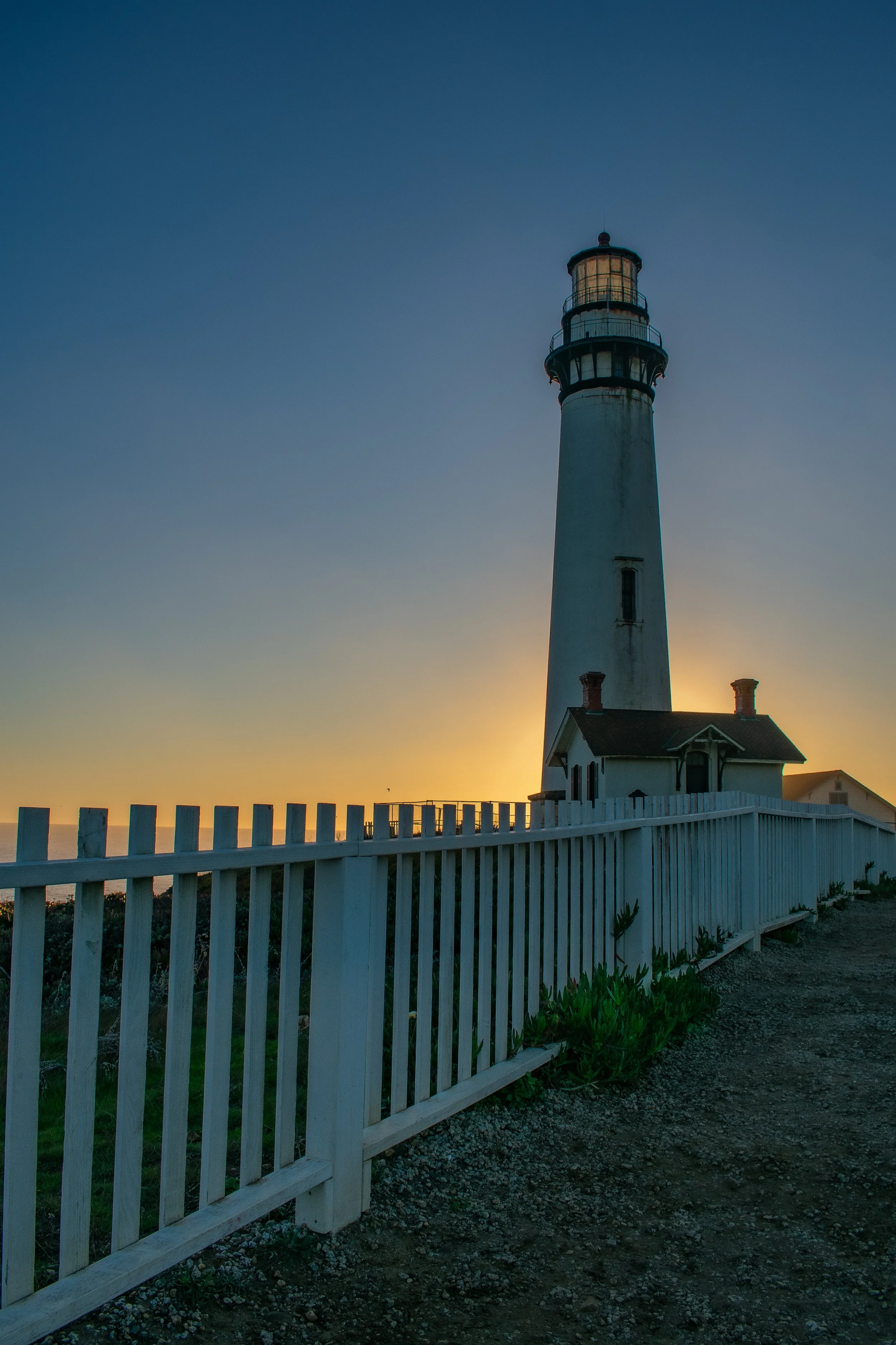 A lighthouse near the coast during sunset with a white picket fence in the foreground.