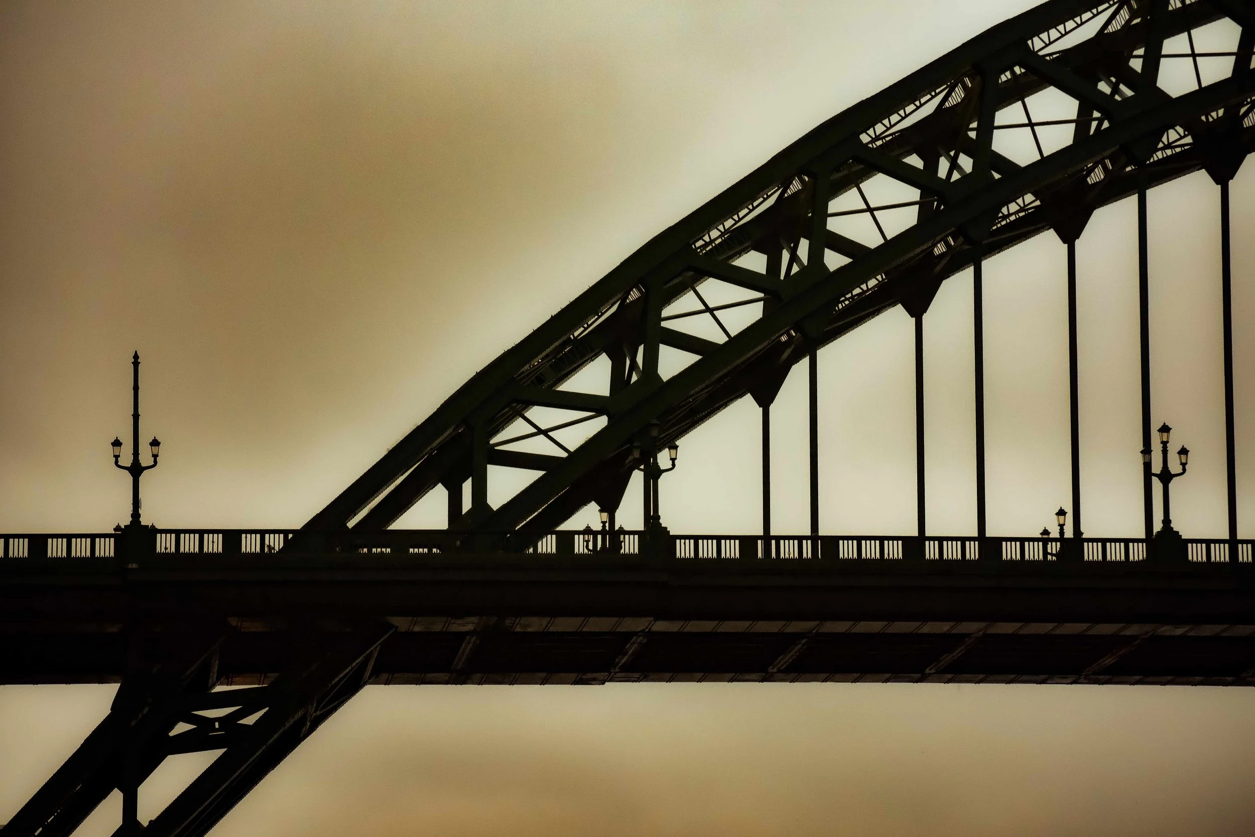 Close-up of a bridge with lamp posts and structural beams against a cloudy sky.