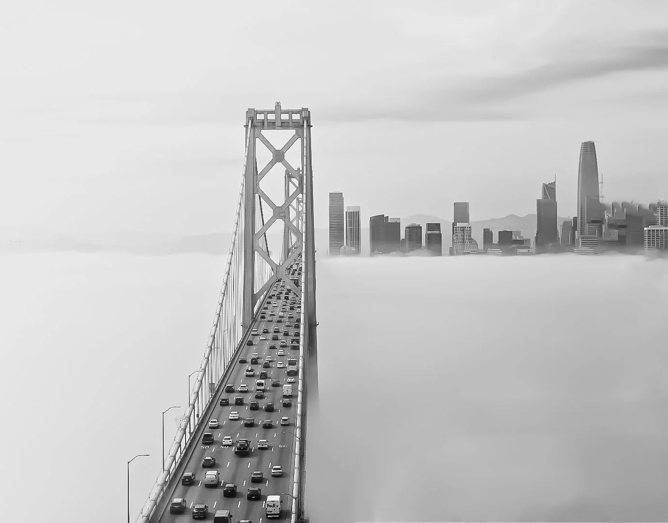 Black and white photo of a foggy San Francisco skyline with the Bay Bridge in the foreground, with numerous cars on the bridge and tall skyscrapers in the background.