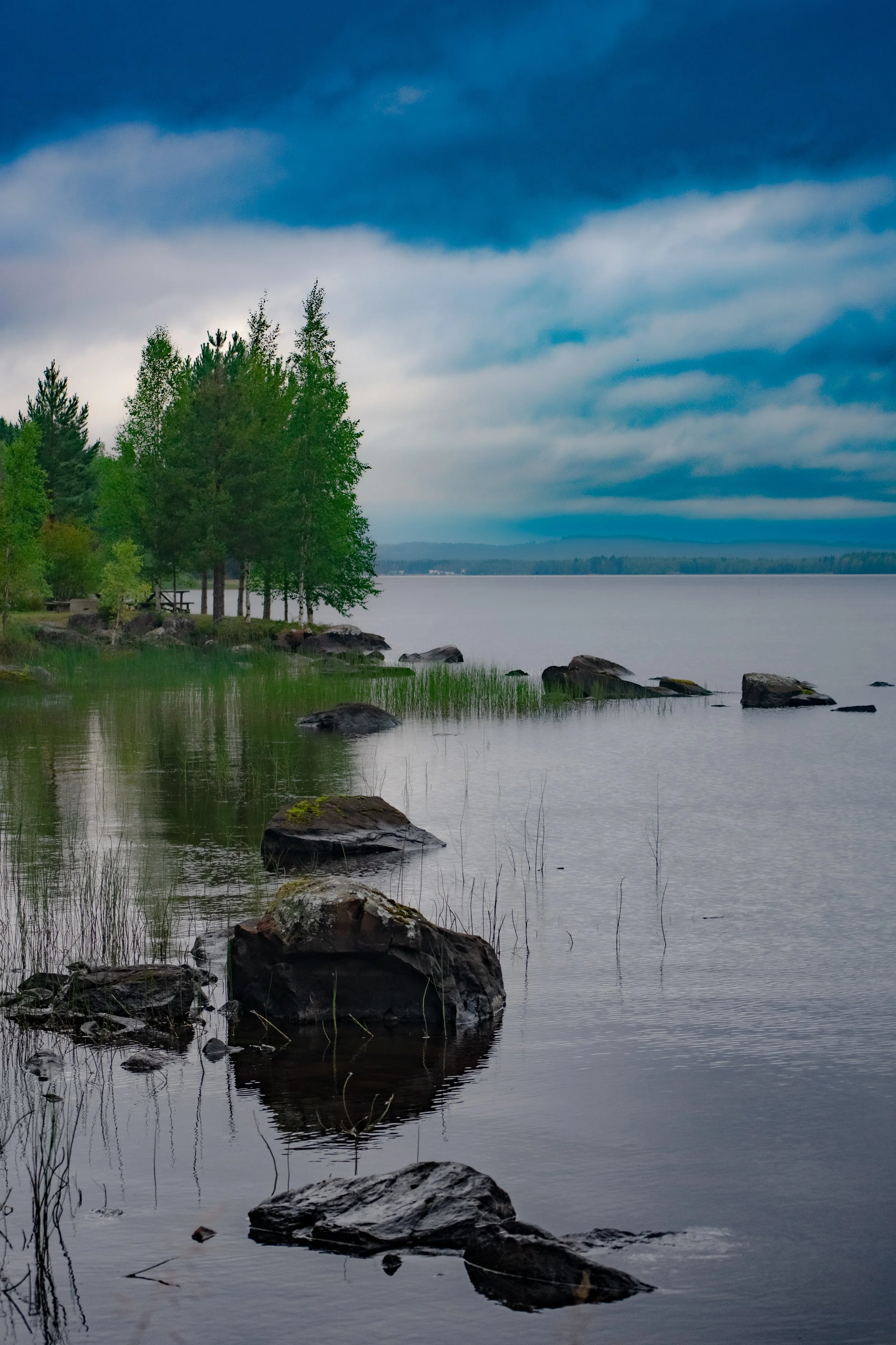 A scenic view of a lake with rocks along the shoreline, green trees, and a cloudy sky.