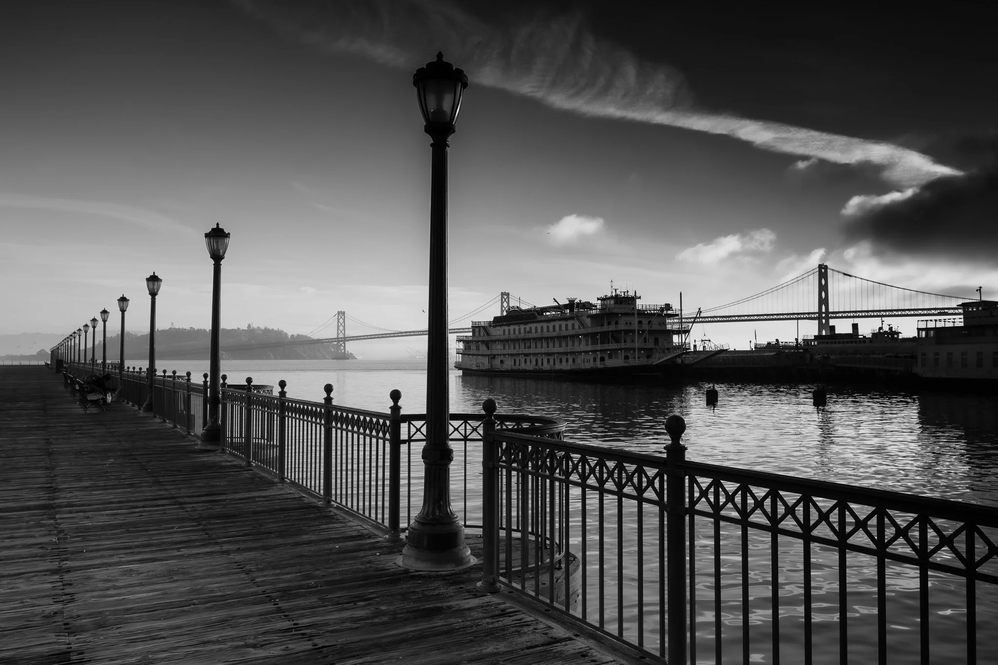 Black and white photo of a wooden pier with a metal railing and street lamps along the water's edge, a large boat docked nearby, a bridge in the background, and cloudy sky overhead.