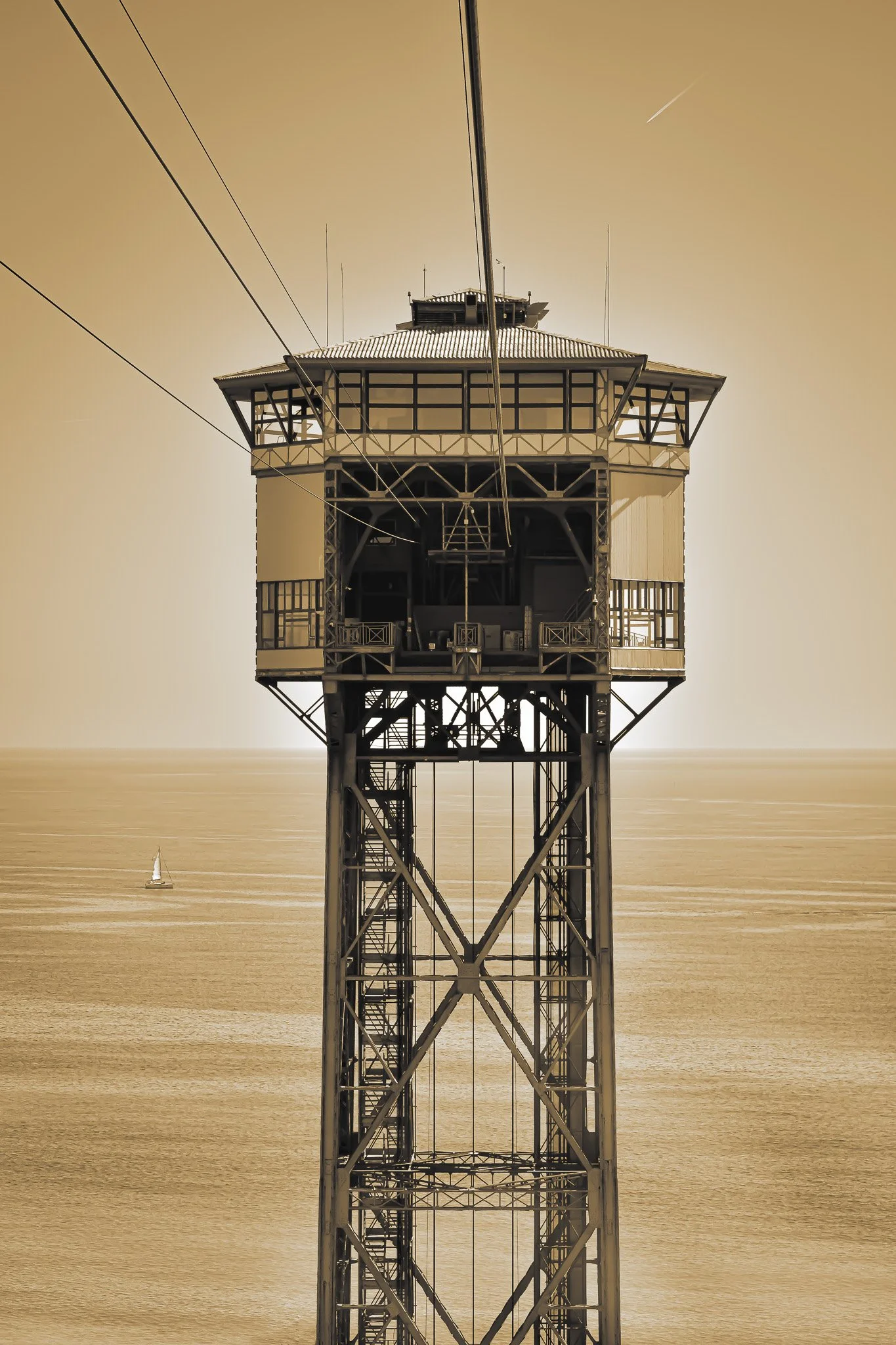 View of a tall, metal, structure-building with a balcony, overlooking a body of water, with a small sailboat and a clear sky in the background.