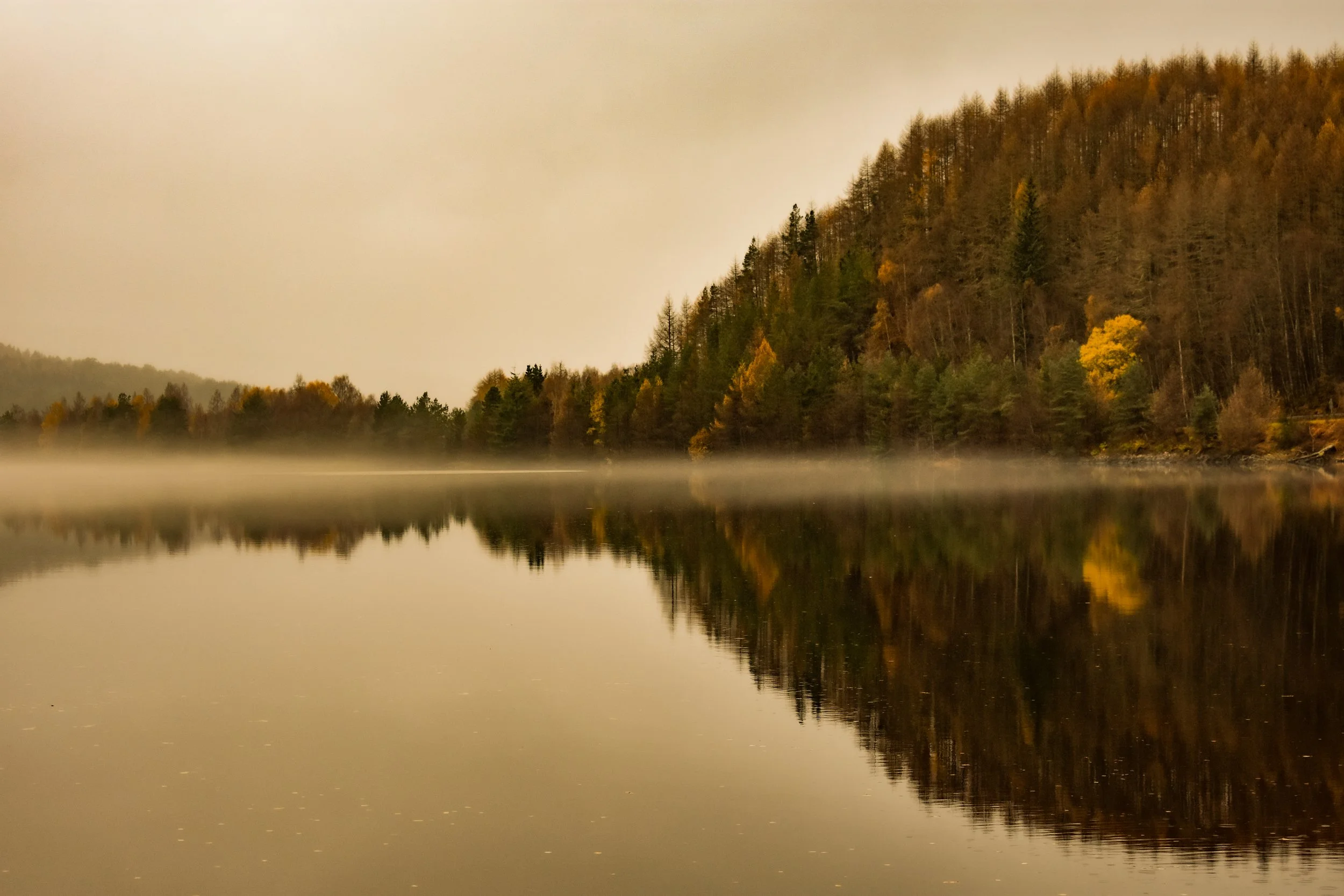 A tranquil lake reflecting a forested hillside with autumn trees, with mist hovering above the water and a cloudy sky overhead.