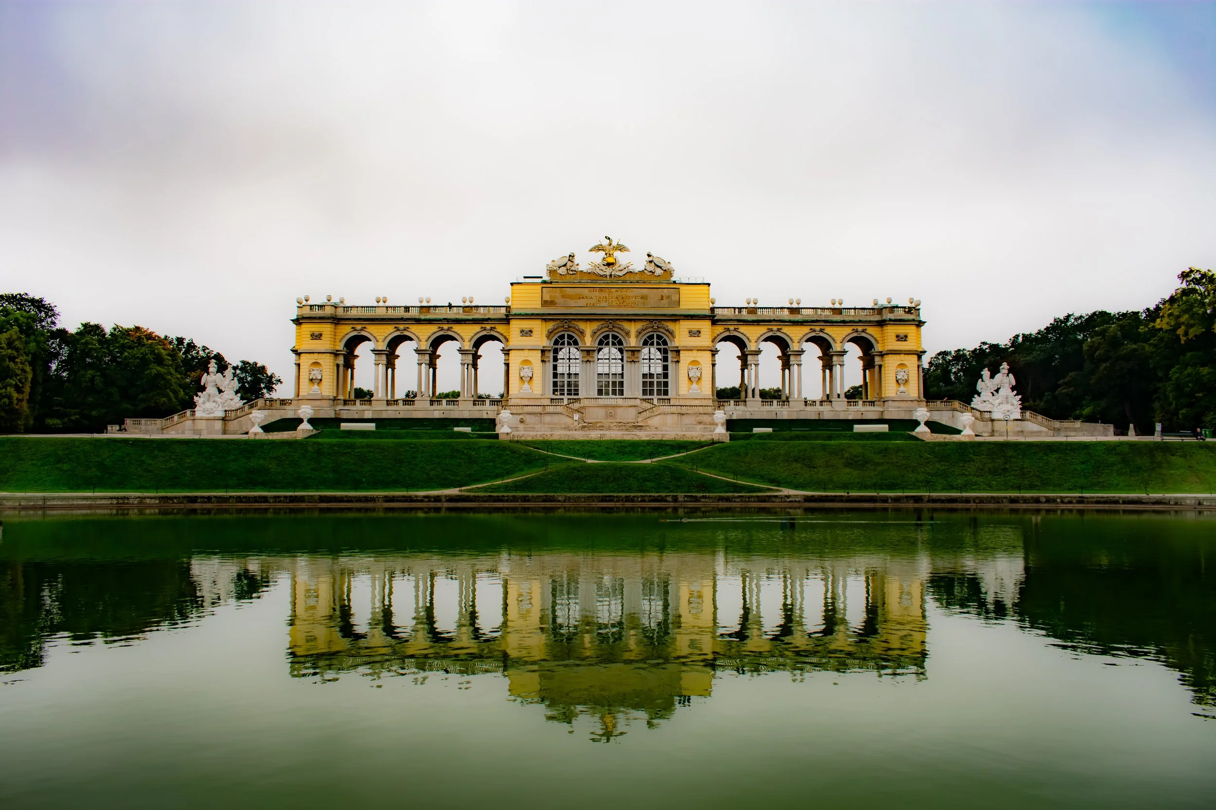 Yellow neoclassical building with arches and sculptures, reflected in a lake, with green trees on either side.
