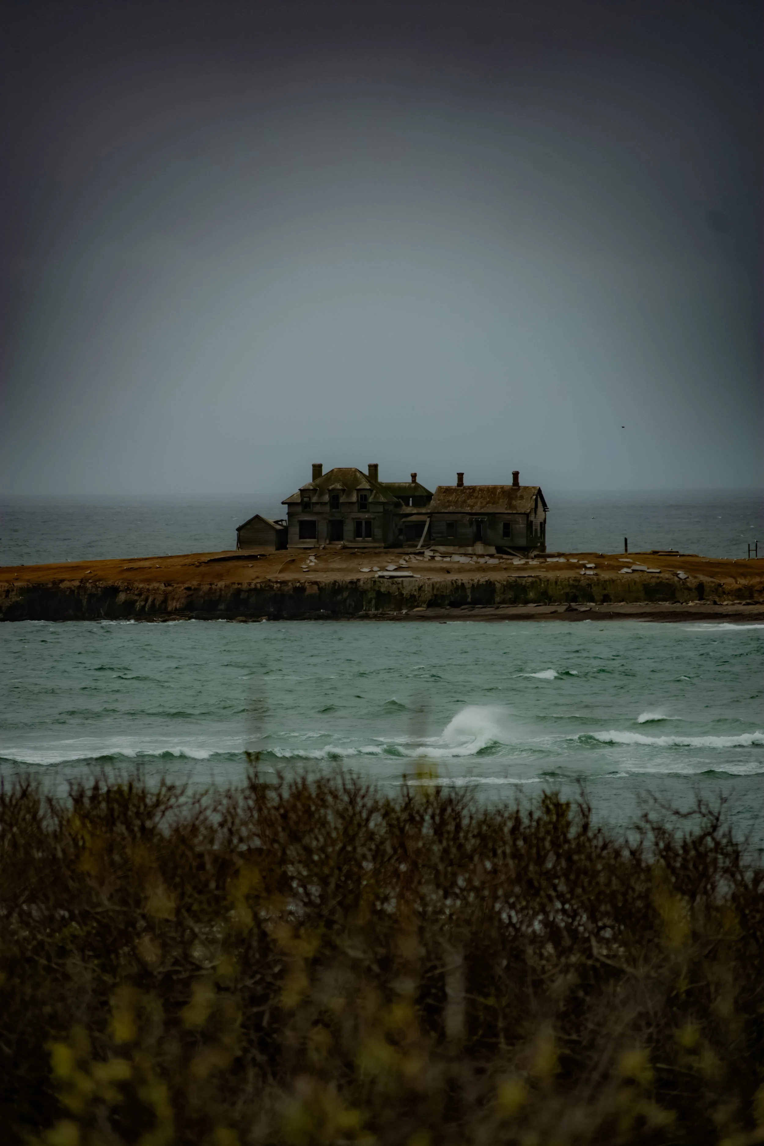 A dilapidated house on a small island, with choppy water and a cloudy, gloomy sky in the background.