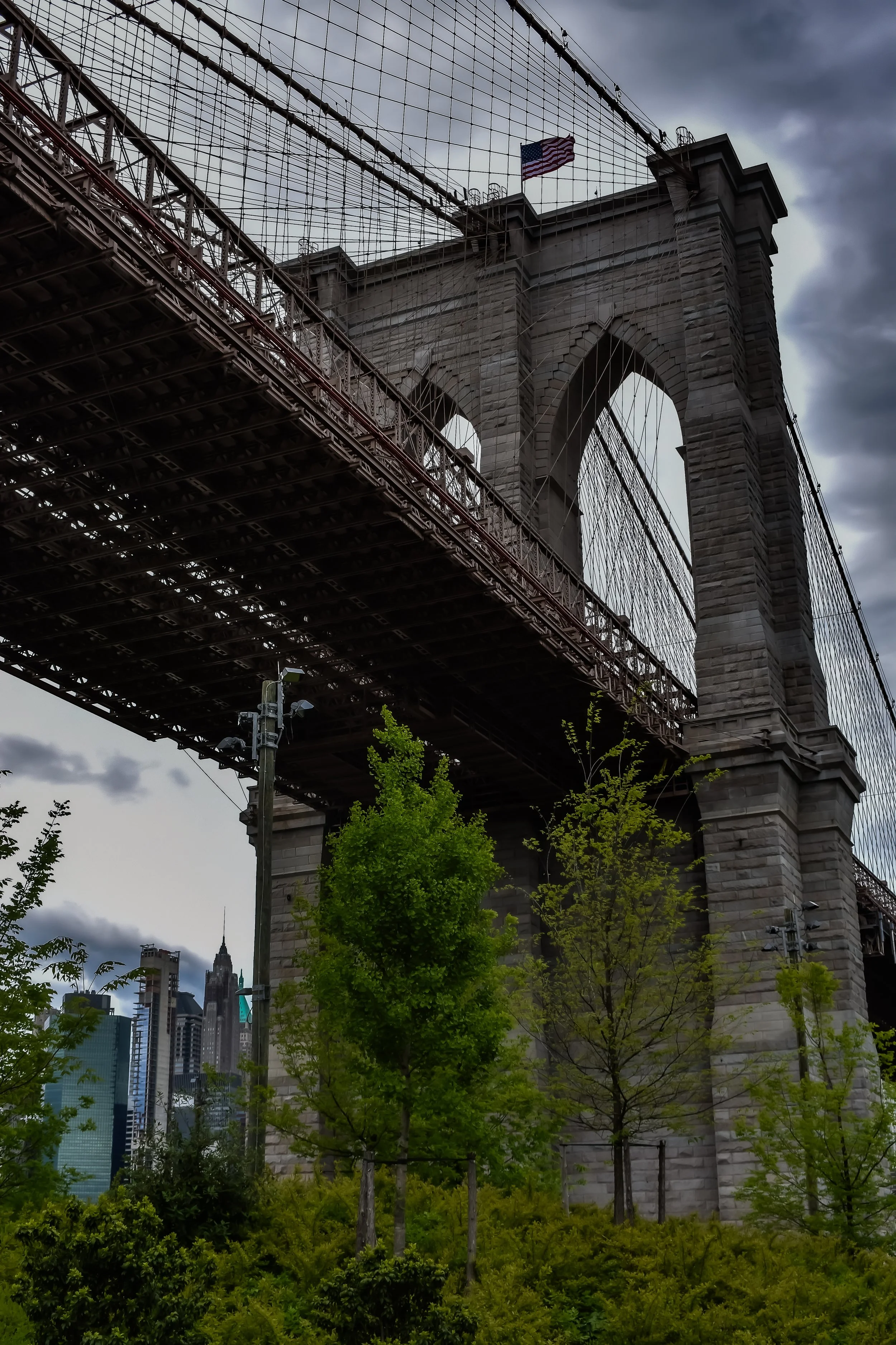 Low-angle view of the Brooklyn Bridge with green trees in the foreground and a cloudy sky in the background.