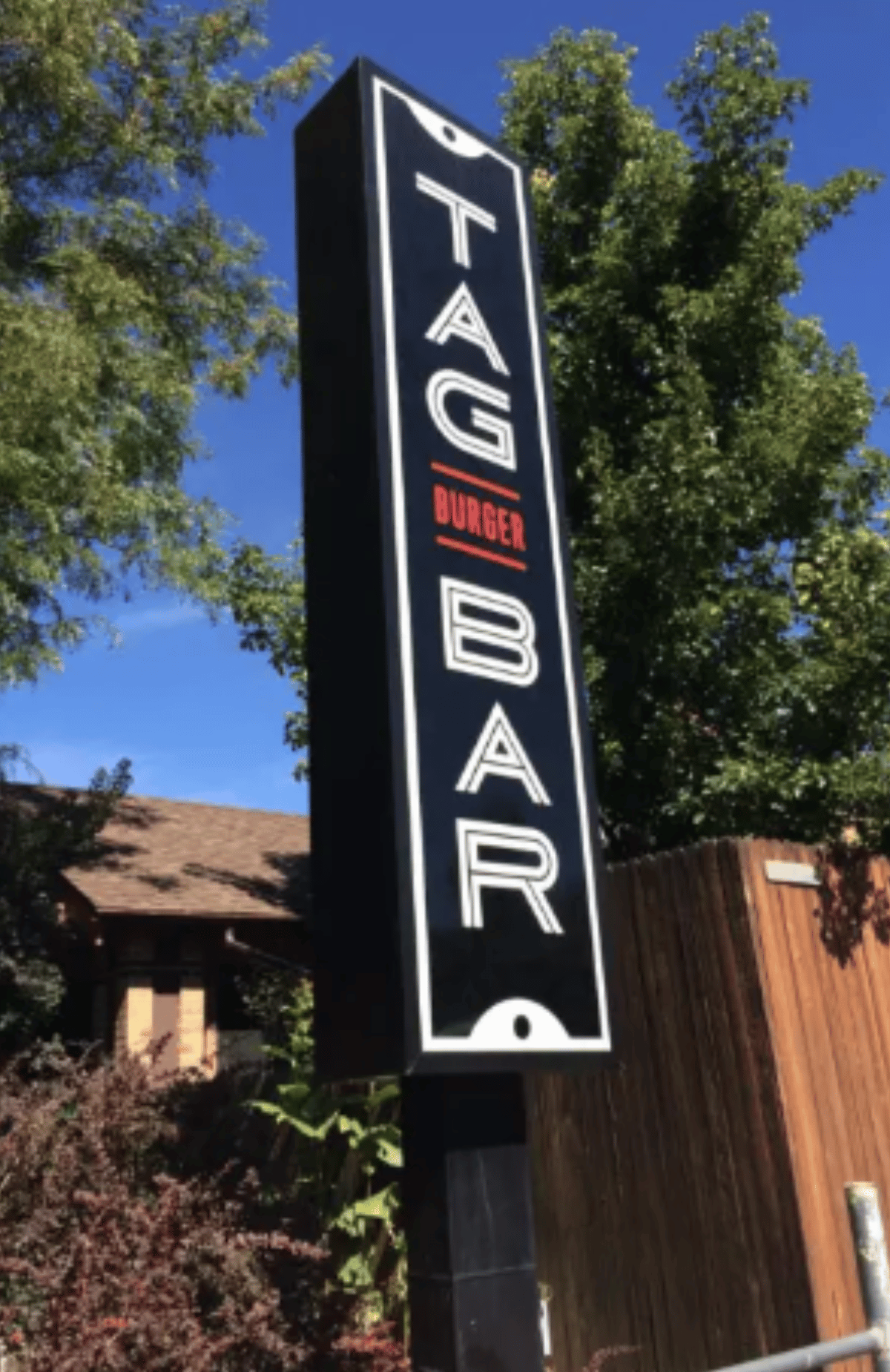Black vertical outdoor sign for Tag Burger Bar in Denver, CO against blue skies and green trees