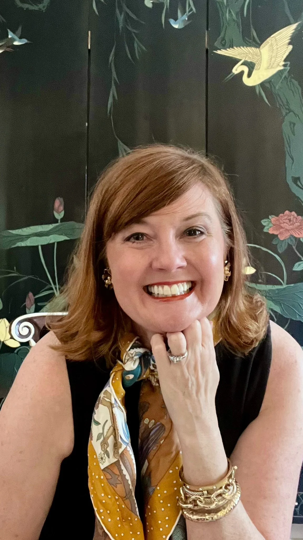 A smiling woman with shoulder-length red hair, gold jewelry, a patterned scarf, and a black sleeveless top, sitting in front of a dark background with artwork of cranes and flowers.
