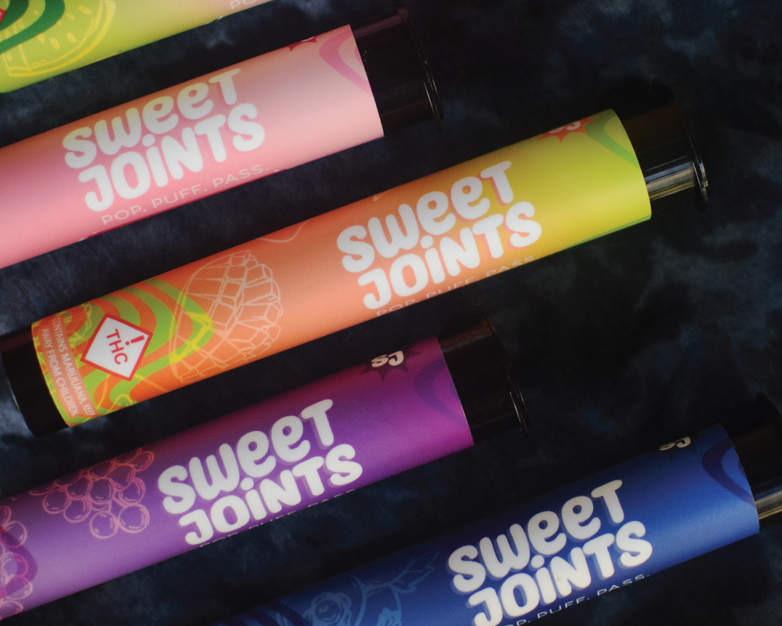 Array of colorful vape pens with the branding 'Sweet Joints' in white, in pink, orange, purple, and blue colors, on a dark fabric background.