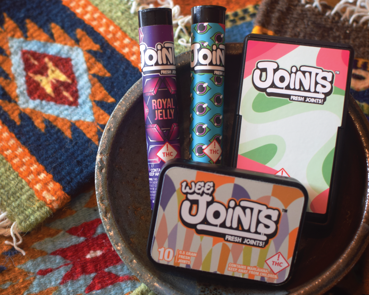 Two THC vape pens with colorful designs, a box of 10 THC joints, and a pack of joints on a rustic plate, with a patterned fabric background.