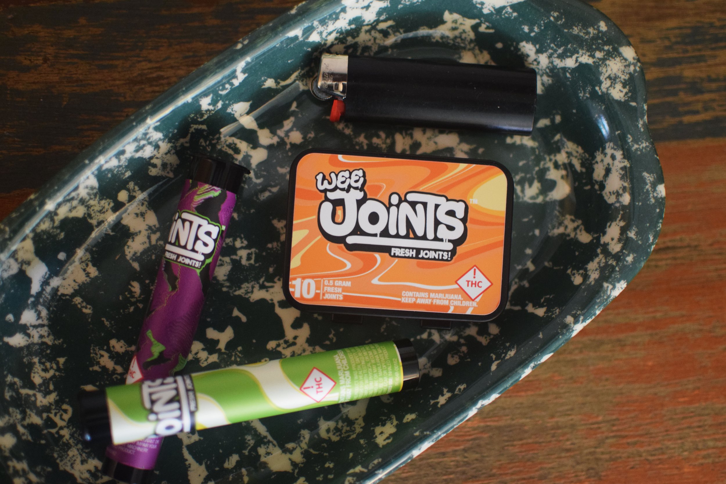 A tin box labeled 'Wee Joints' with cannabis joints and a lighter on a chipped green surface.