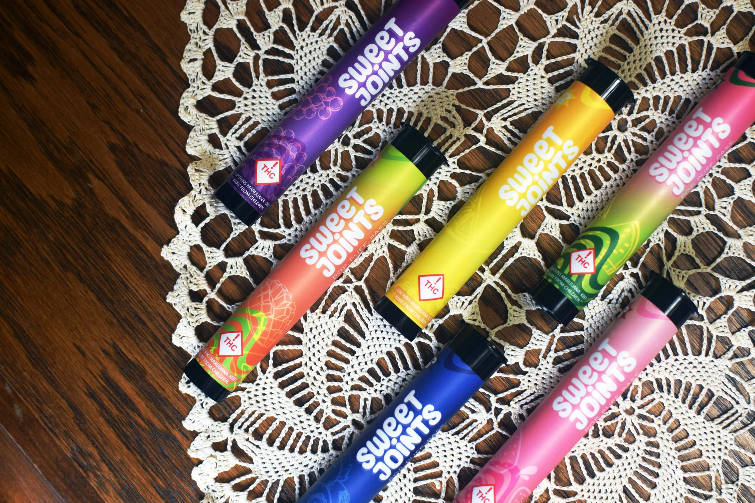 Five colorful THC-infused vape pens on a lace doily on a wooden surface, with colors purple, orange, yellow, green, blue, and pink, labeled 'Sweet Joints'.