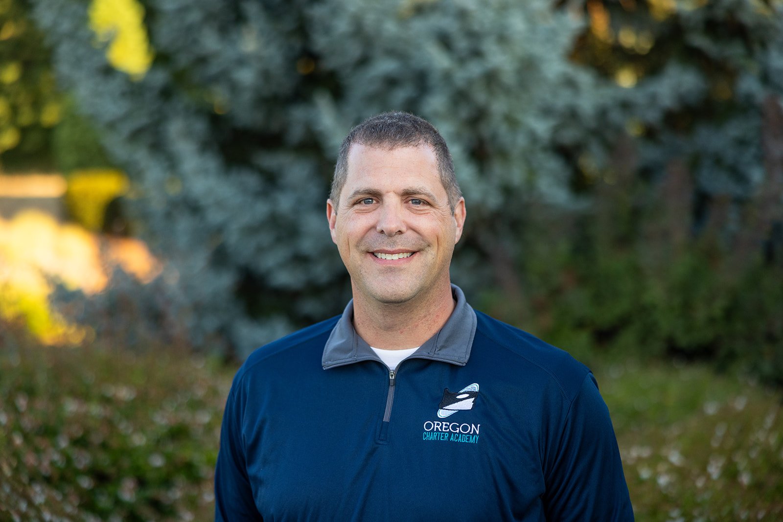 A man wearing a navy blue Oregon Charter Academy polo shirt standing outdoors with trees and plants in the background, smiling at the camera.