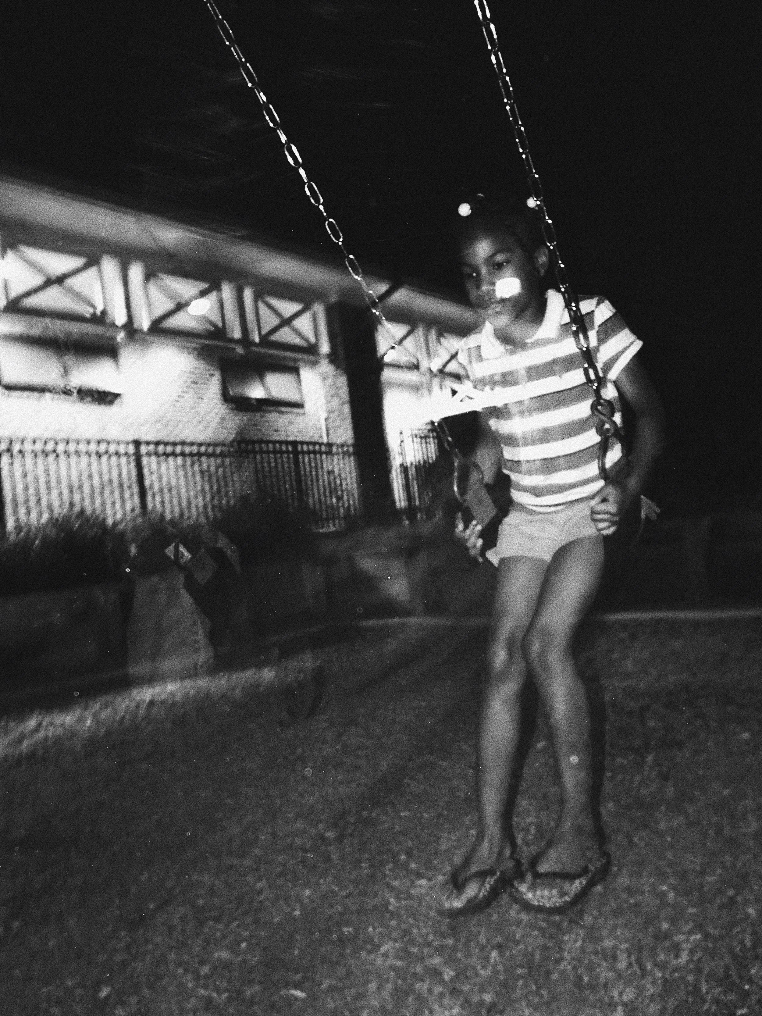 Black and white photo of a child on a swing at night, wearing a striped shirt and shorts, with a building in the background.