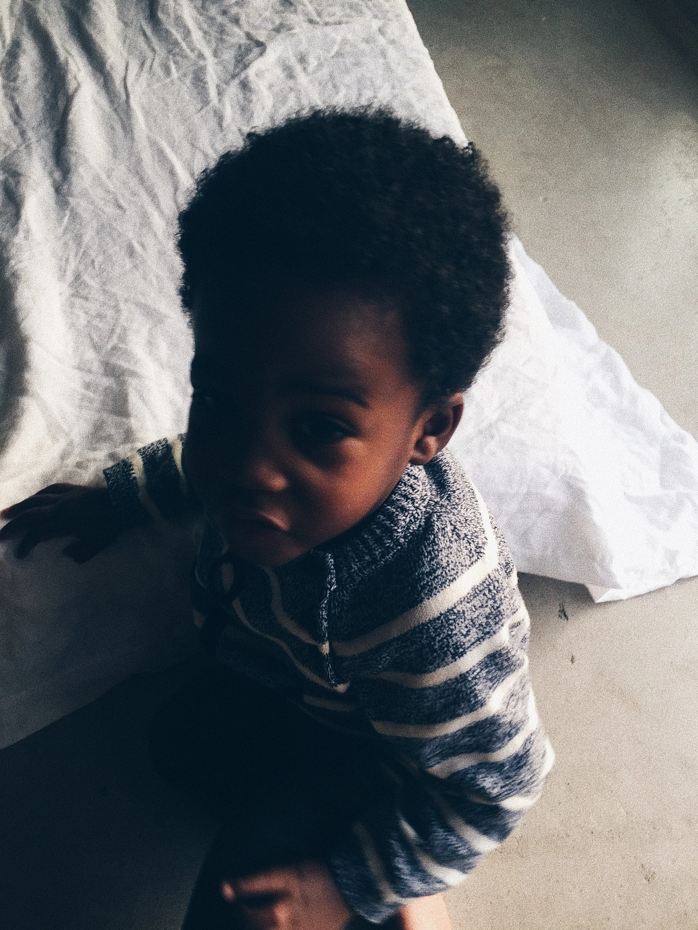 Young child sitting on a floor, wearing a striped sweater, next to a crumpled white sheet.