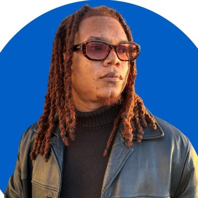 Person with long locs and sunglasses wearing a leather jacket against a blue background.