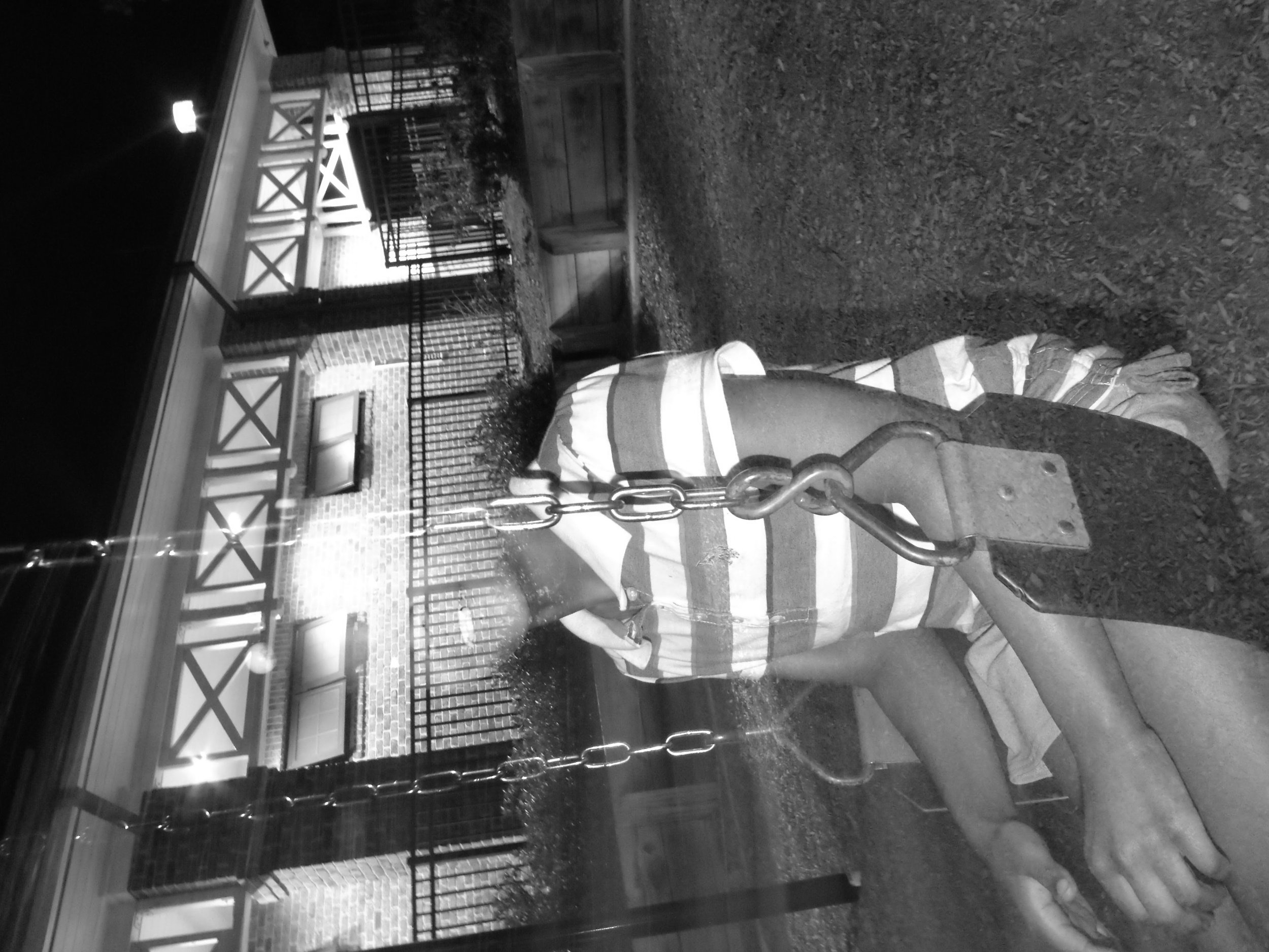 Black and white photo of a person on a swing at night, with a brick building in the background.