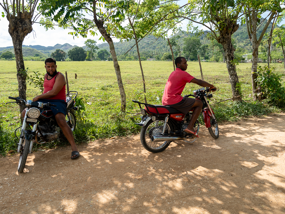 Two people on motorcycles parked on a dirt road beside a grassy field, surrounded by trees, with hills in the background.