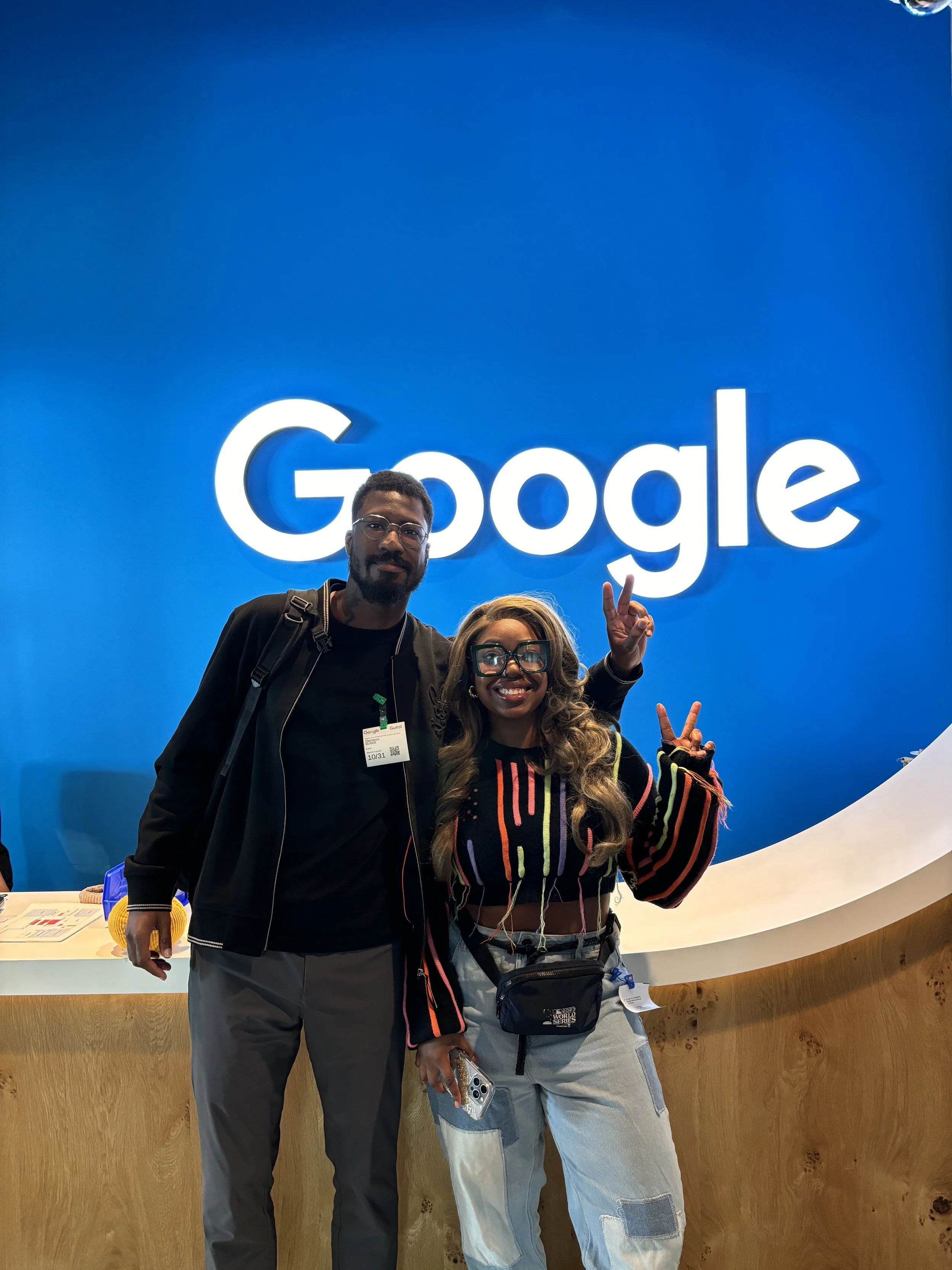 Two people standing in front of a Google logo on a blue wall, smiling and posing for the camera. One person is making a peace sign, and they are both casually dressed. The counter in front of them is wooden.