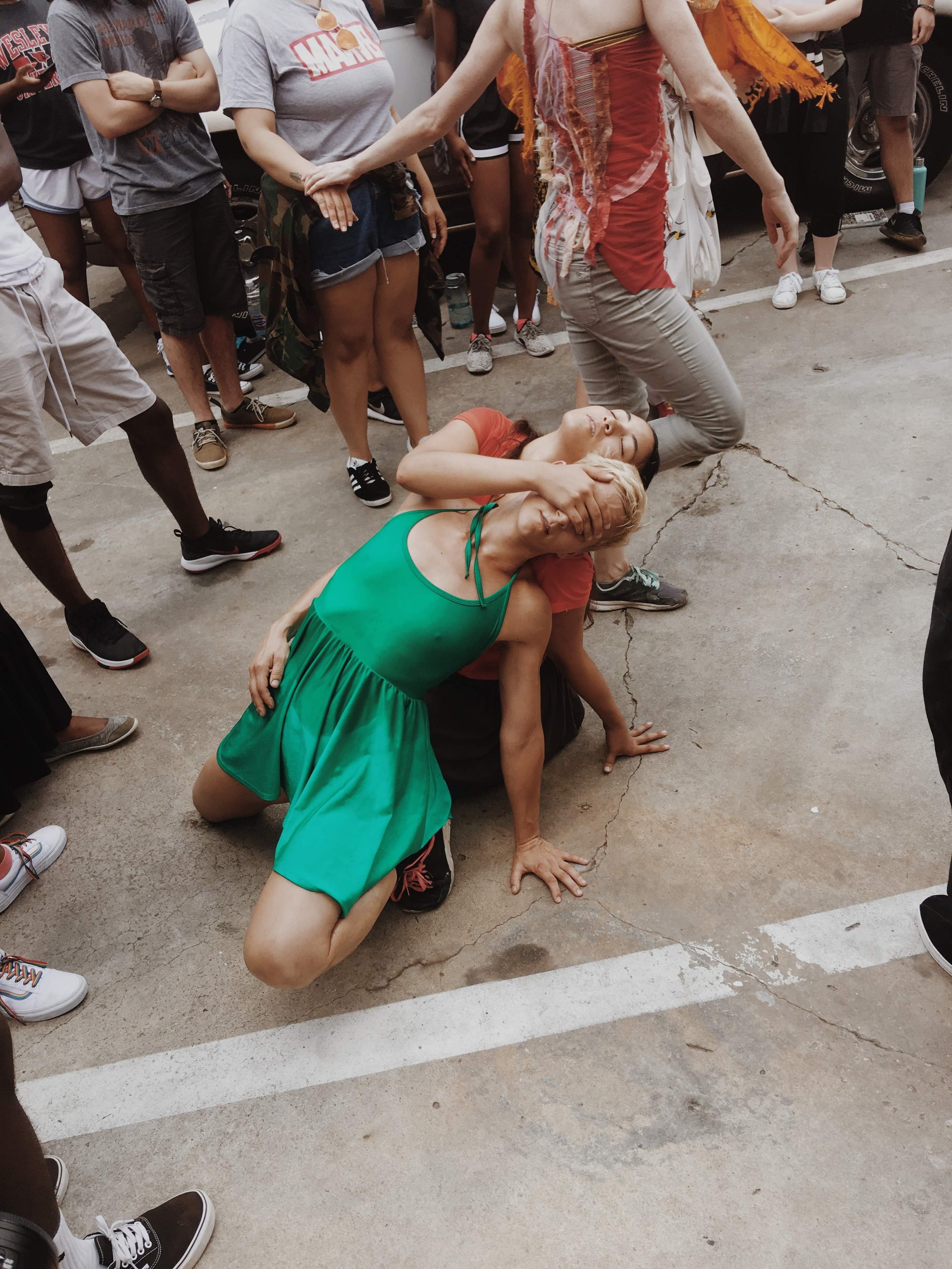 Two performers in an outdoor street performance, with one in a green dress leaning on the other, surrounded by a standing crowd.