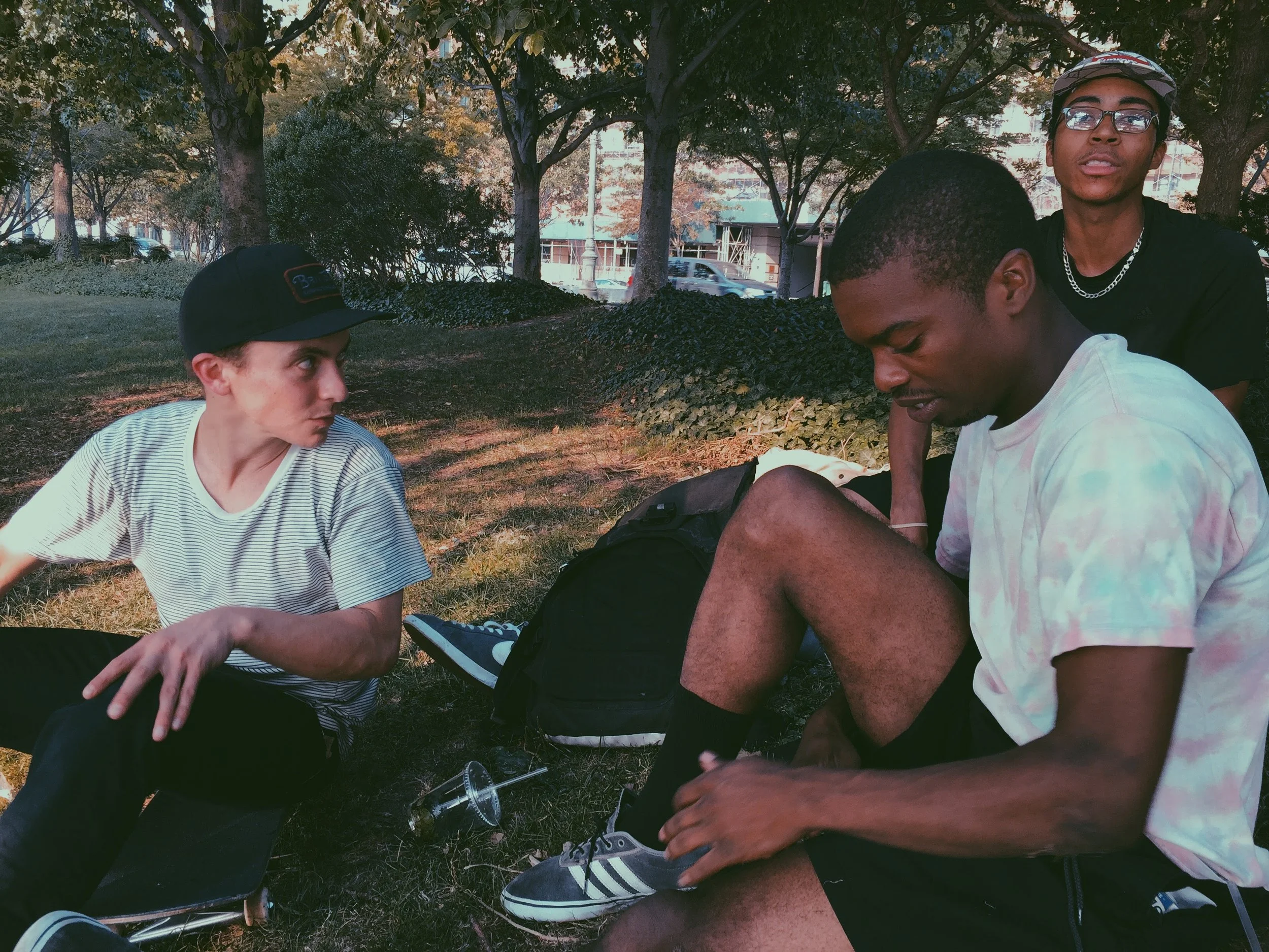 Three young men sitting on the grass in a park, one wearing a striped shirt and cap, another tying his shoe, and the third standing behind. Skateboards and a drink are nearby.
