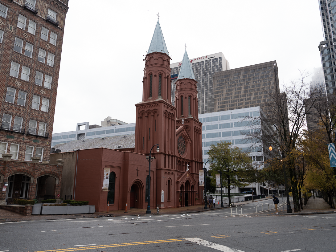 Historic red brick church with two spires and a circular stained glass window, surrounded by modern city buildings and an empty street.