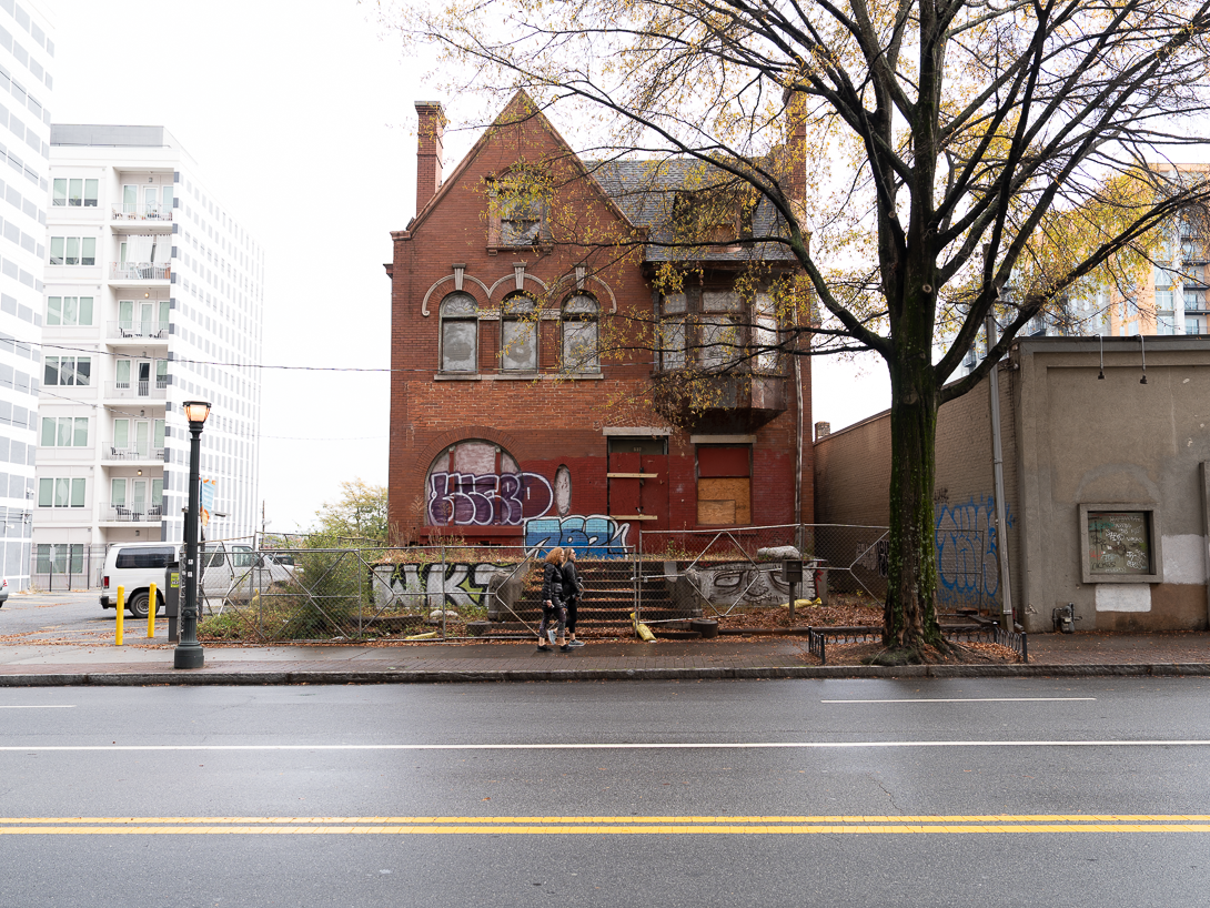 An abandoned brick building with boarded-up windows and graffiti, located next to a tree on an urban street. Newer buildings and a parked van are visible in the background. The scene includes a wet sidewalk and street, indicating recent rain, and a couple of pedestrians walking by.