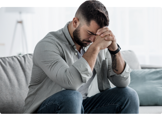 A man sitting on a sofa with his head bowed and hands clasped in front of his face, appearing upset or stressed.