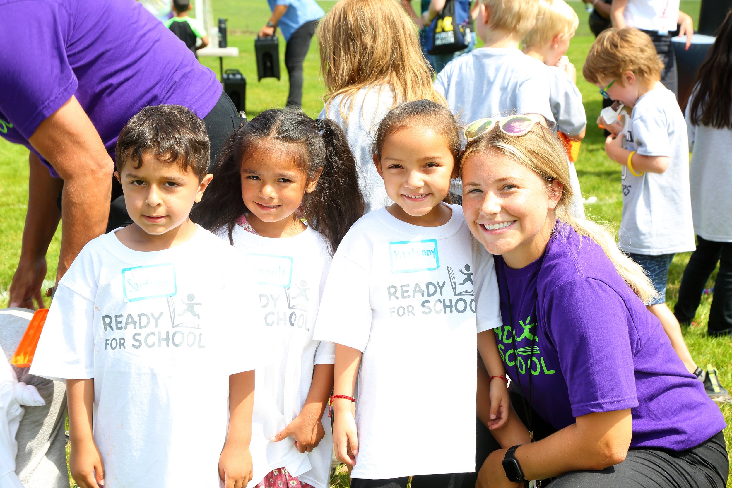 A group of smiling children wearing white t-shirts with 'Ready for School' printed on them, standing outdoors on a grassy field during a school event. An adult woman, smiling and wearing a purple t-shirt, is squatting next to the children.