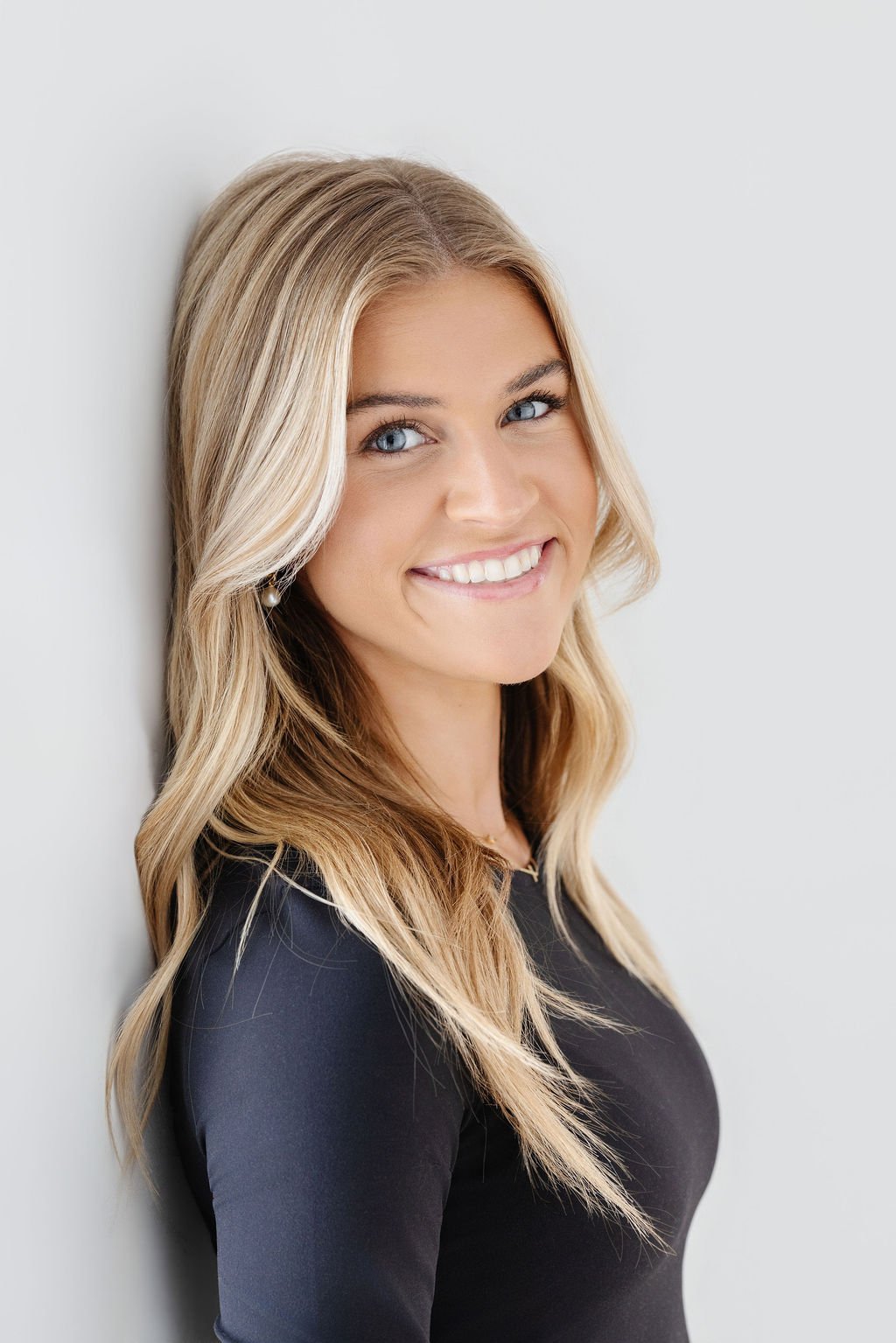 Portrait of a smiling woman with long blonde hair, wearing a black shirt, standing against a white background.