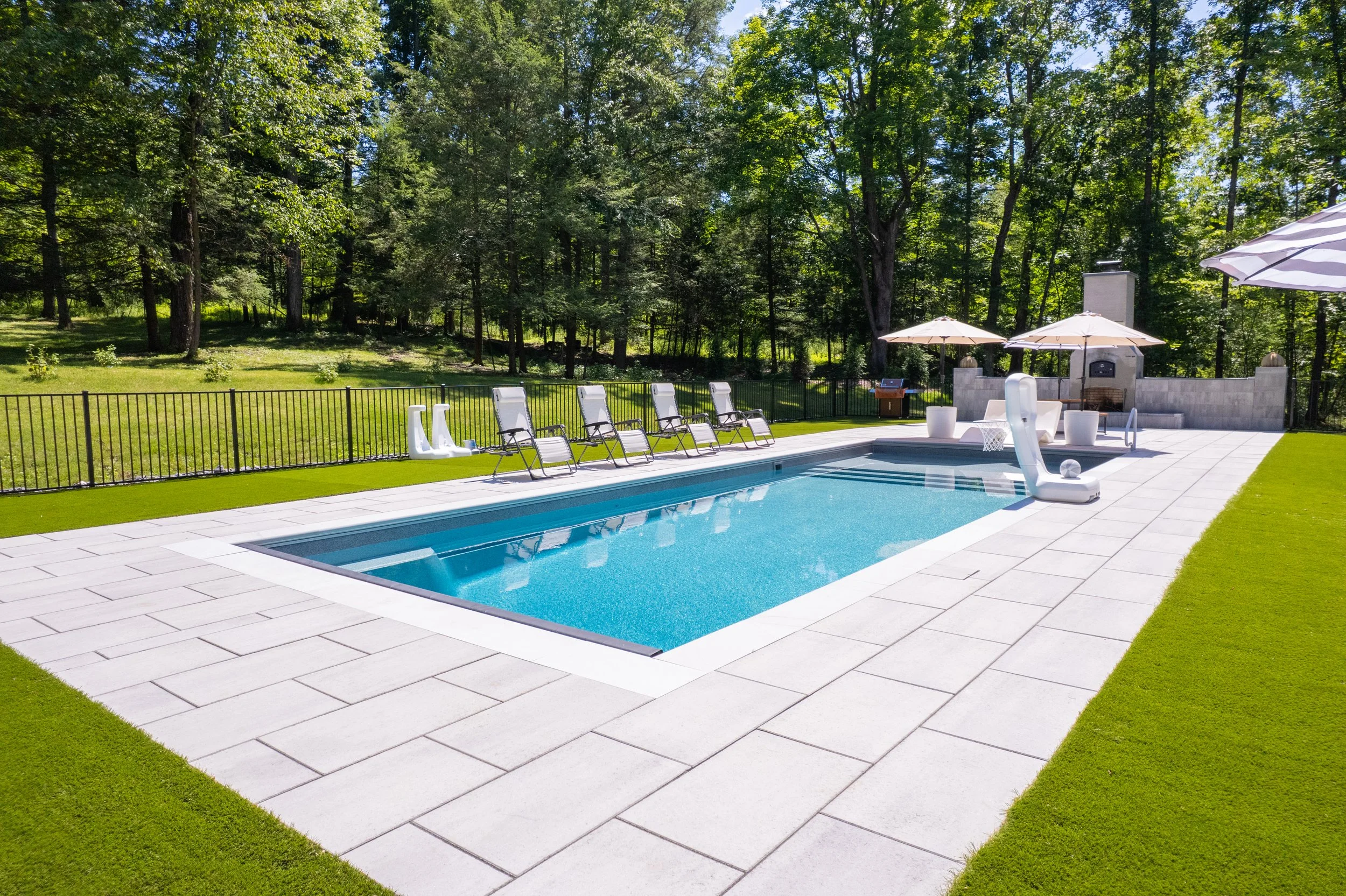 Daytime backyard pool with white patio, lounge chairs, and umbrellas.