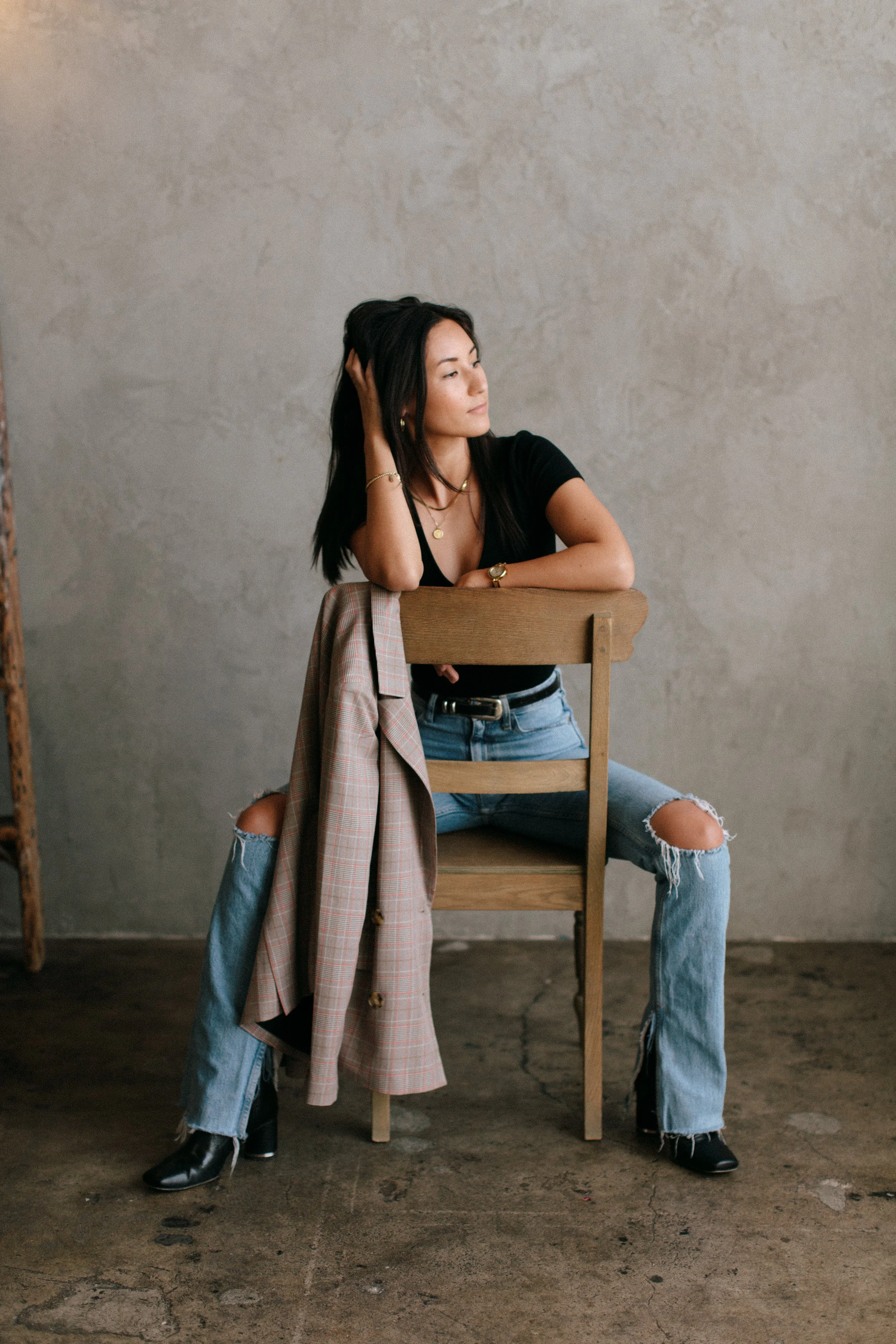 A woman with long dark hair sitting on a wooden chair in front of a plain textured wall, wearing a black top, ripped jeans, and black boots, with a plaid jacket hanging on the chair.