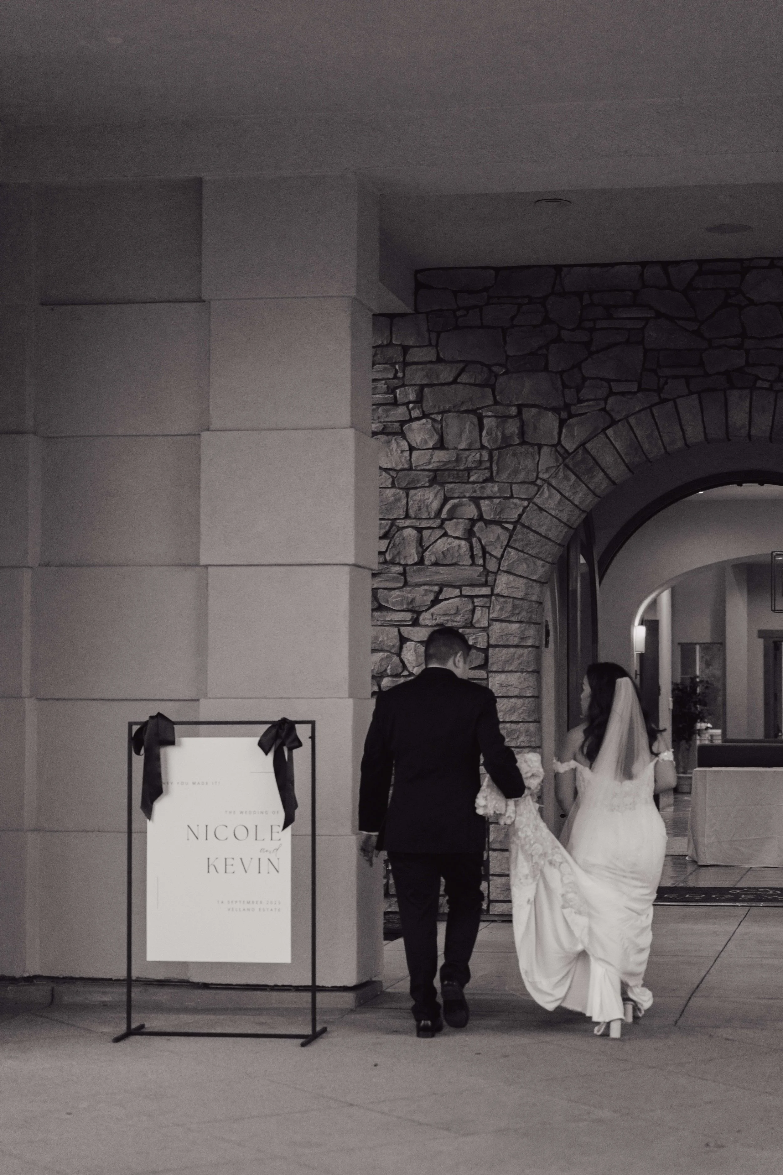A bride and groom holding hands, walking into a wedding venue with stone and brick architecture. The bride is wearing a white wedding gown and veil, and the groom is dressed in a black suit. There is a sign on a stand with ribbons that say 'Nicole and Kevin' and the wedding date, September 2023.