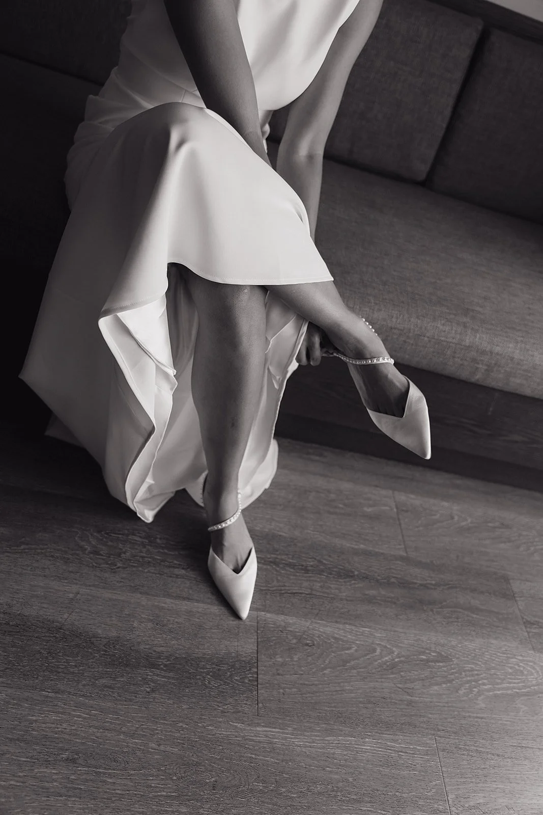 A woman in a white dress is sitting on a couch, putting on high-heeled shoes, with one leg crossed over the other.