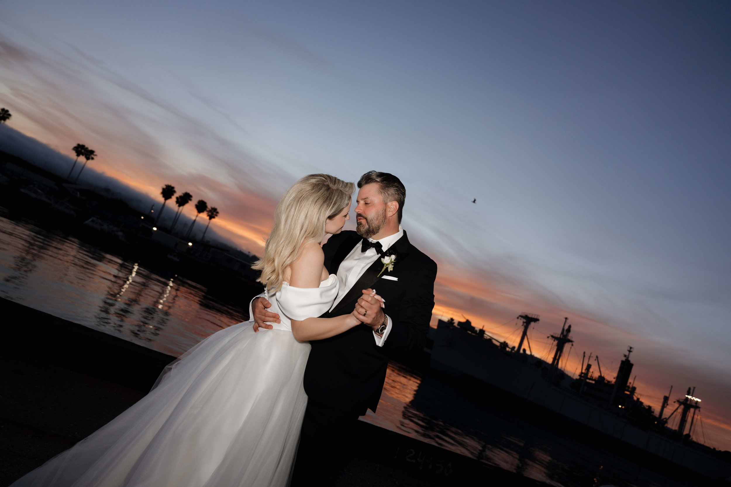 A bride and groom dancing outdoors during sunset, with palm trees, water, and industrial structures in the background.