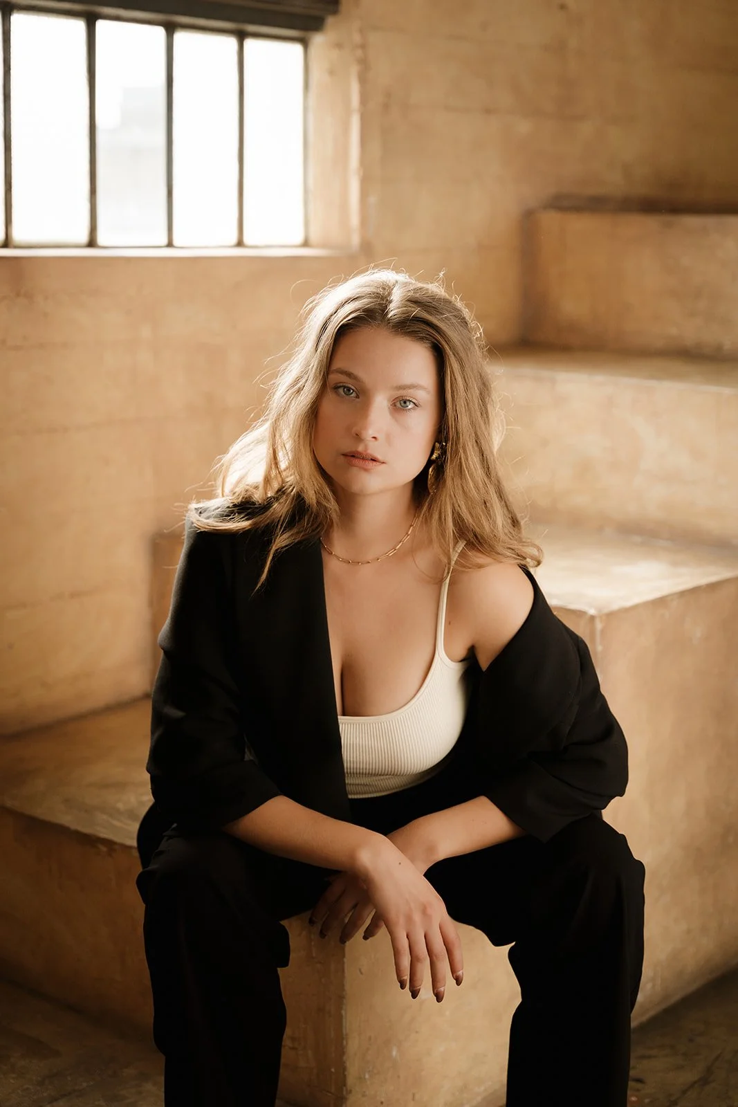 A young woman with wavy blonde hair sitting on a beige concrete staircase near a window, wearing a black blazer, a cream-colored tank top, and black pants.